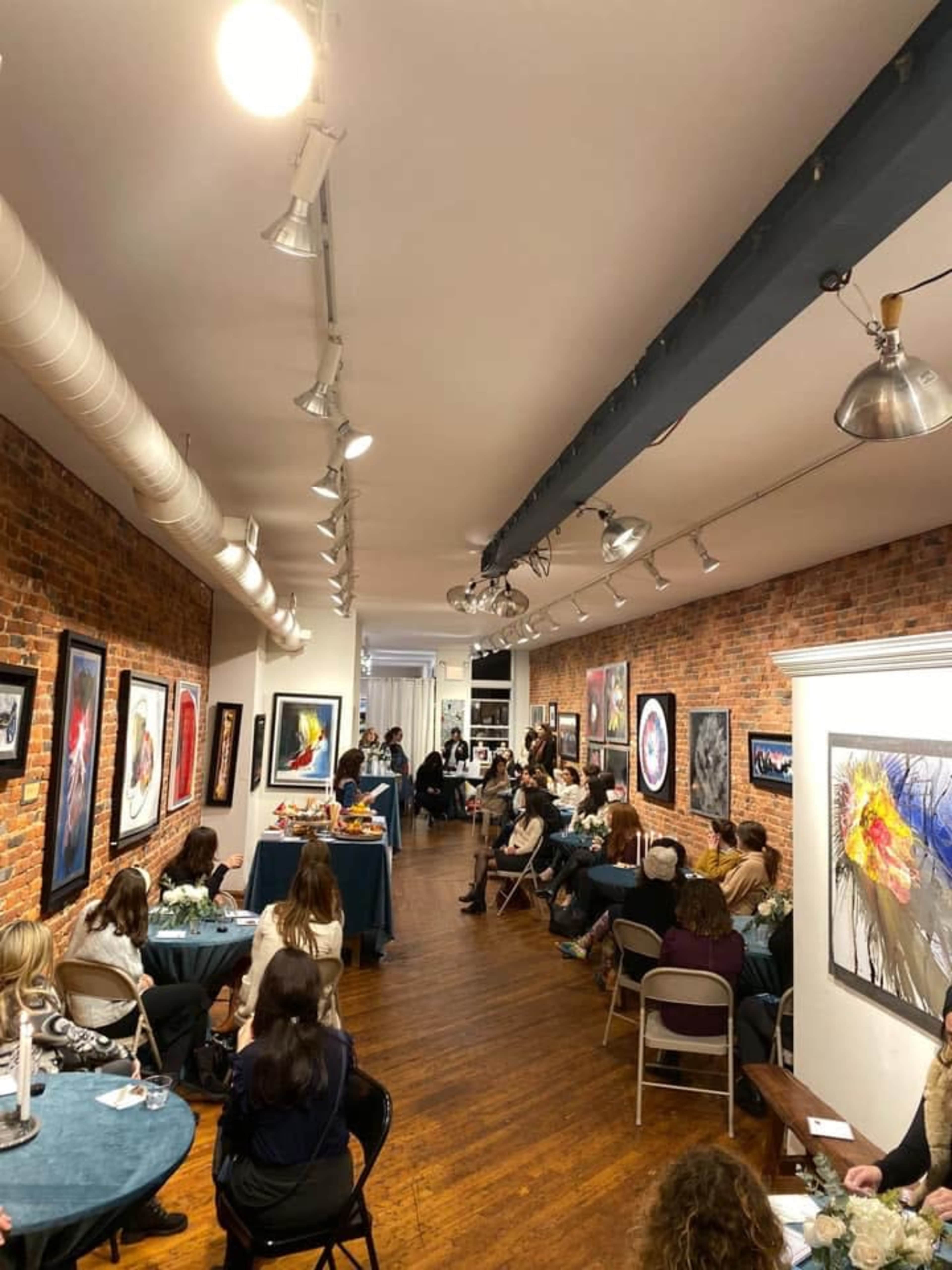 A group of people sits in a gallery with exposed brick walls, surrounded by various framed artworks, while some attendees listen to a speaker at the front.