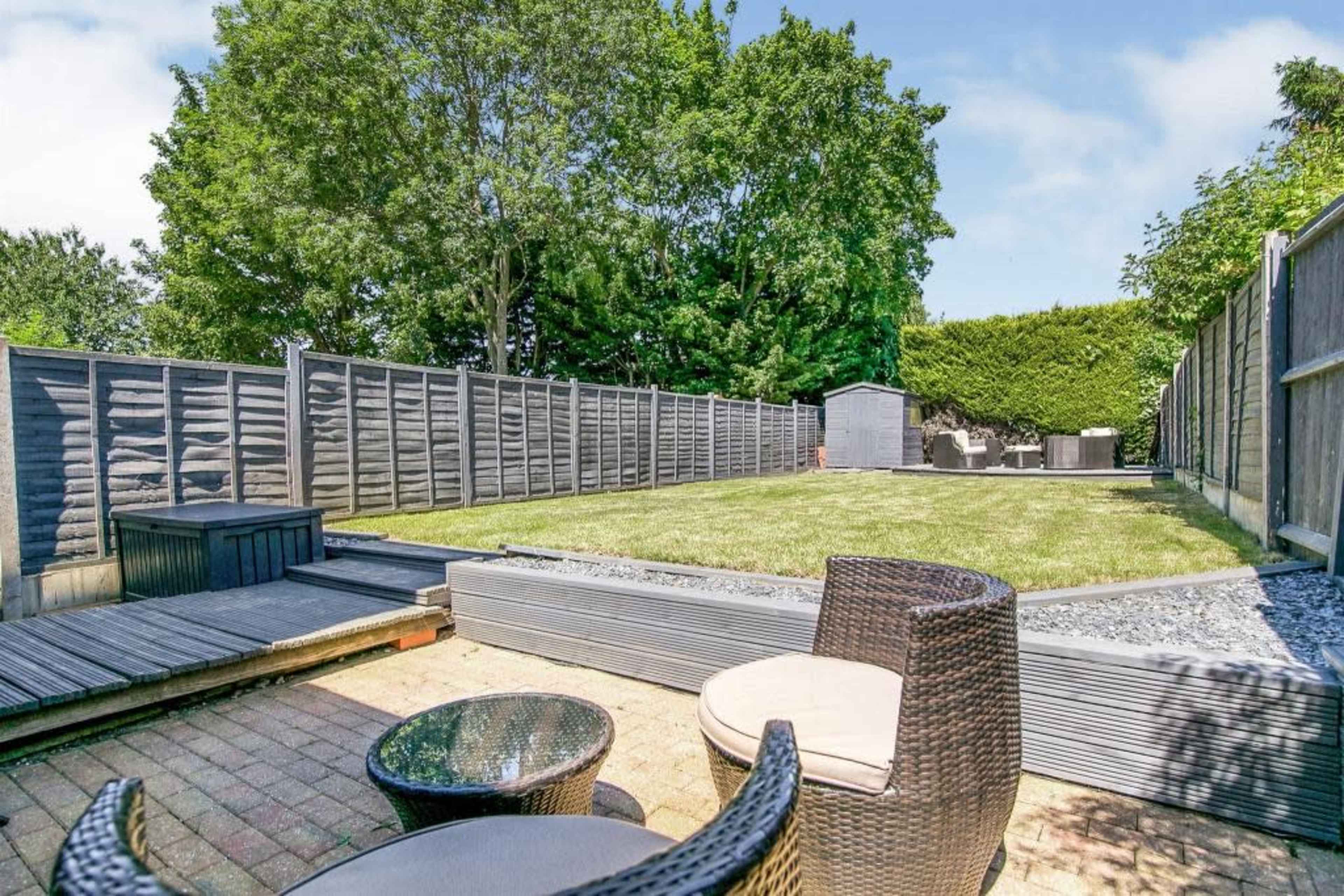 A landscaped garden with a grassy area, a wooden deck, and a stone pathway, surrounded by wooden fences and trees.