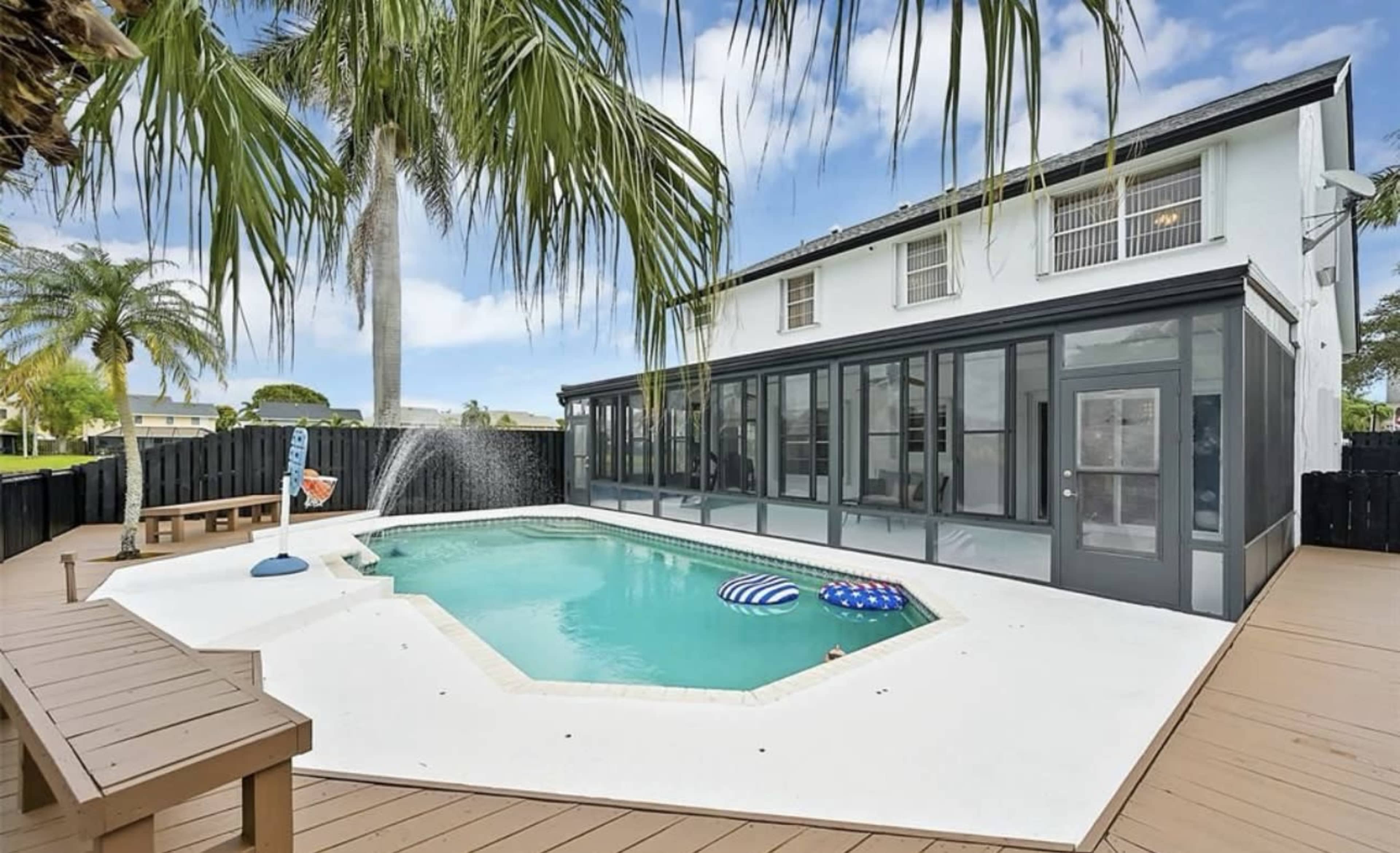 The image shows a backyard with a swimming pool surrounded by a wooden deck and a two-story house in the background.