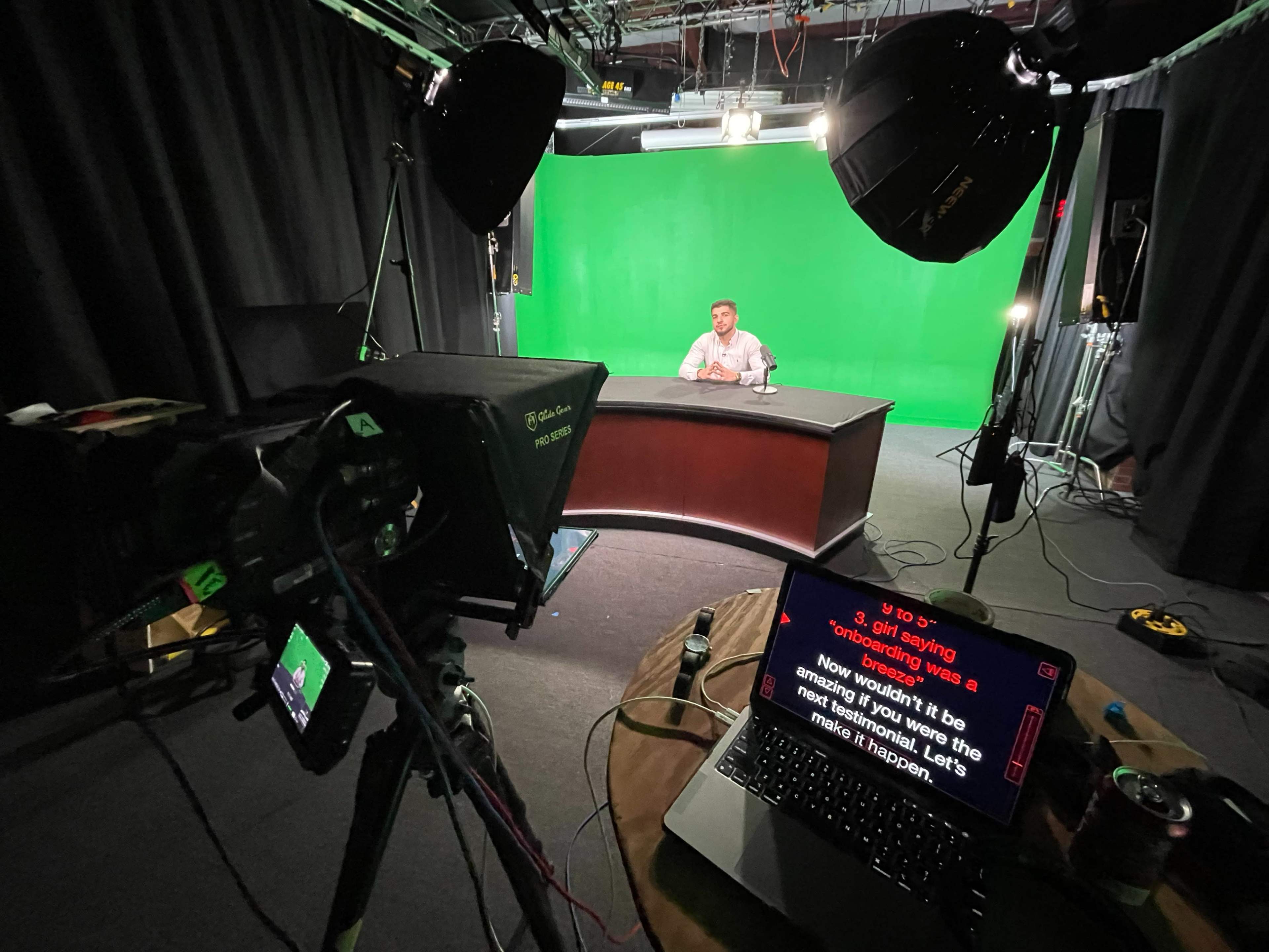 A person is sitting at a desk in front of a green screen, with studio lights and a teleprompter visible in the foreground.