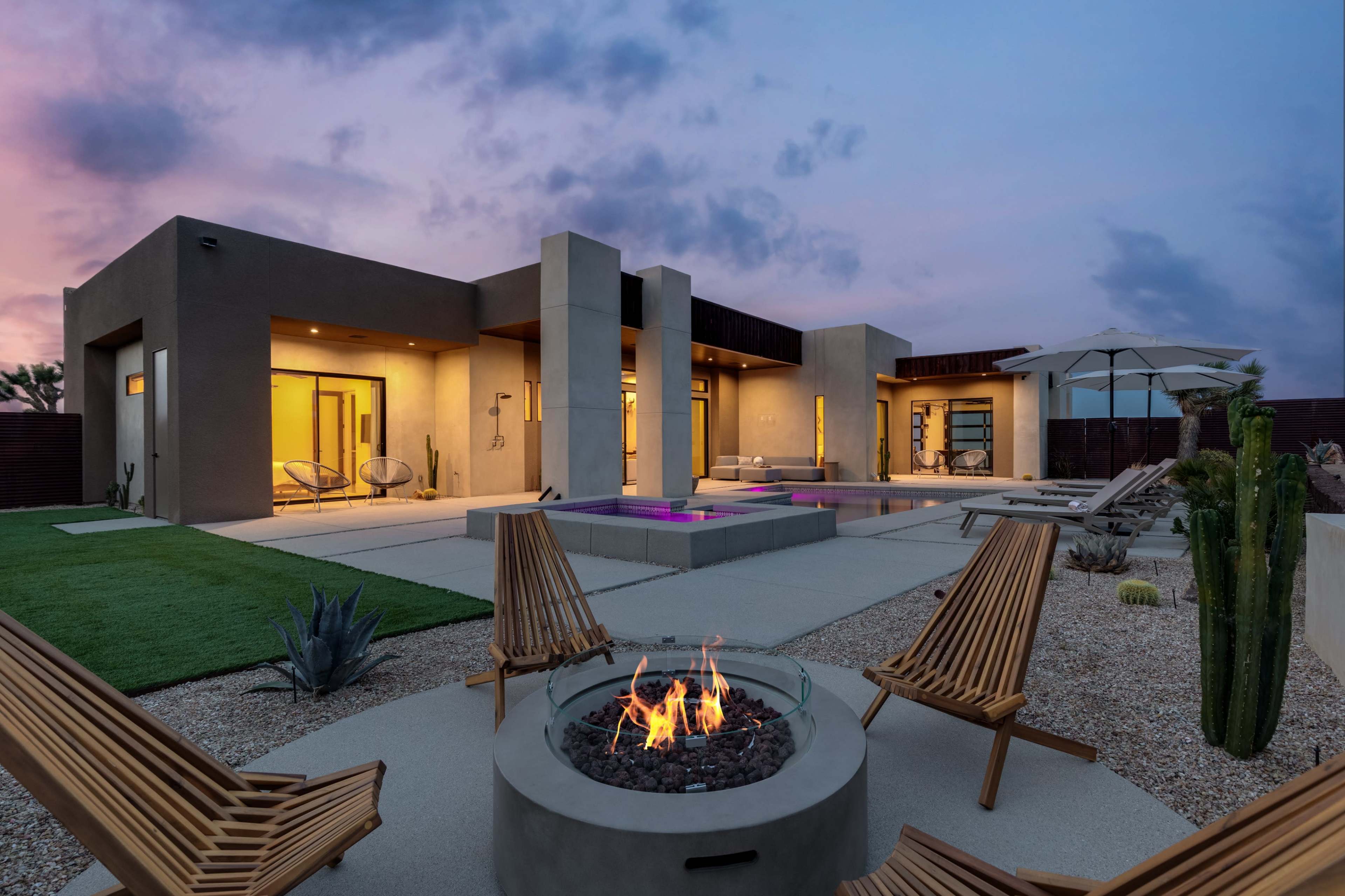 A modern home with large windows and a backyard featuring a fire pit, lounge chairs, and a pool, set against a colorful sunset sky.