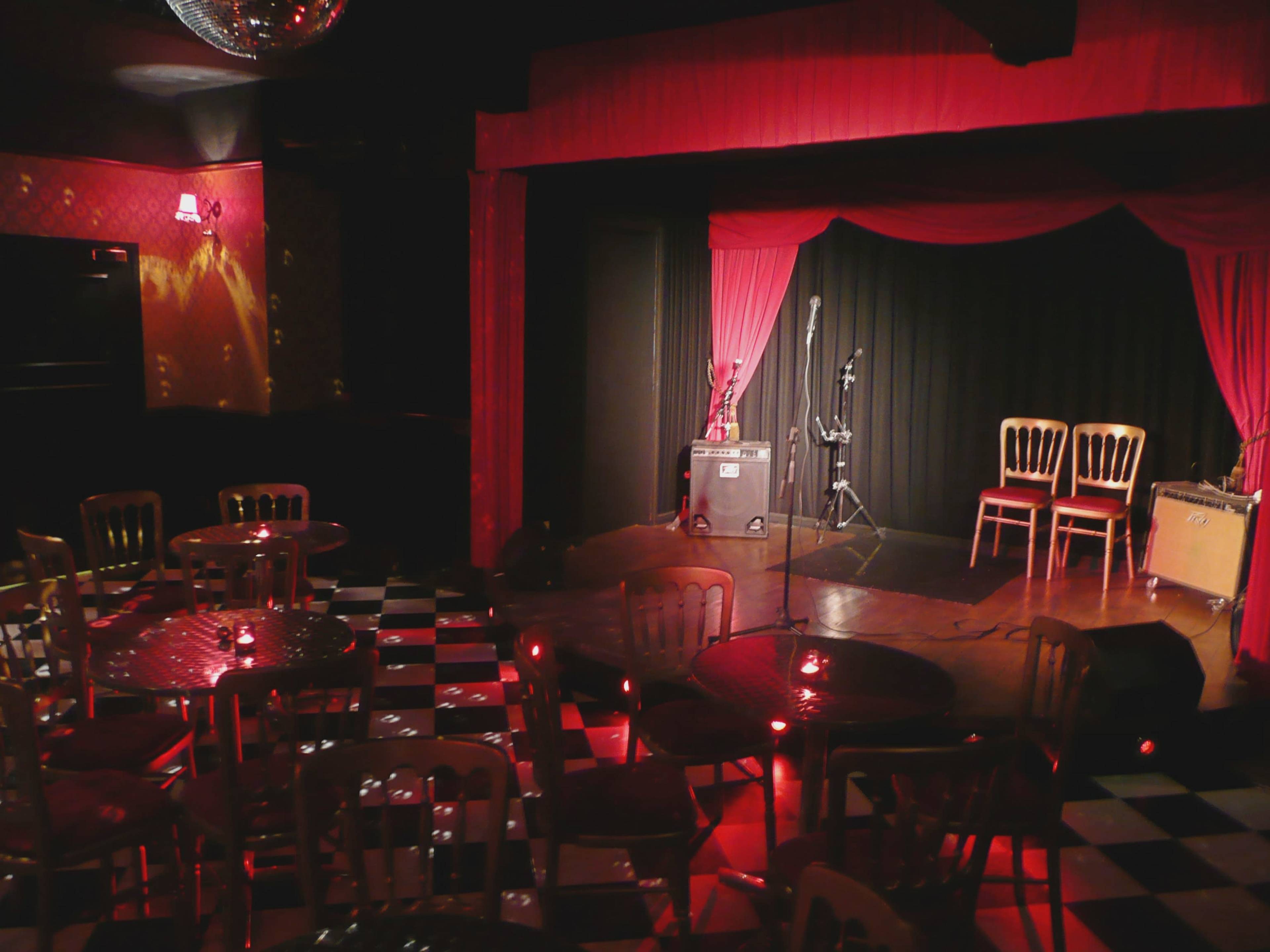 A small music venue with a stage featuring red curtains, two microphones, and three empty chairs, surrounded by tables and chairs on a checkered floor.