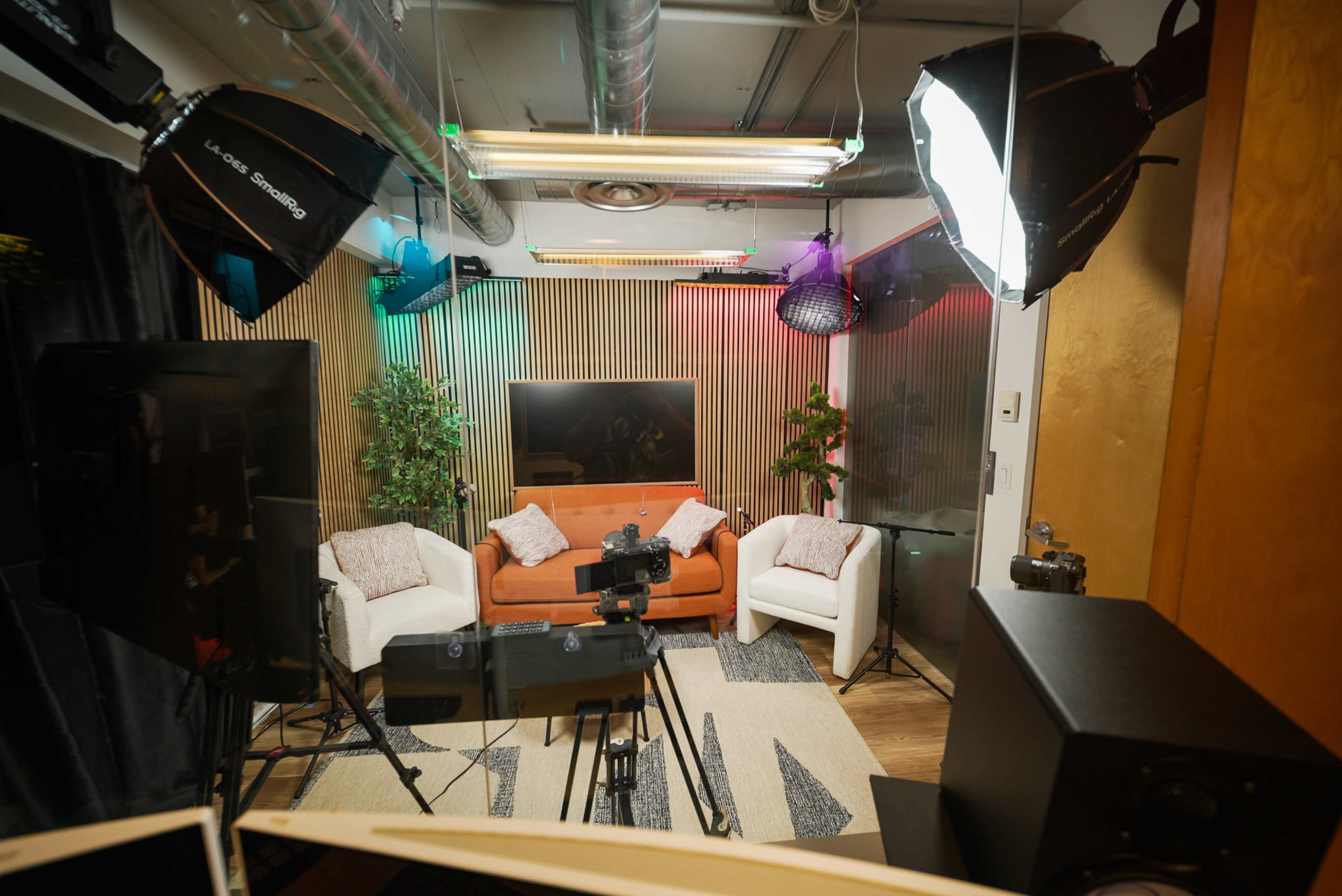 The image shows a modern studio setup with a colorful backdrop featuring a couch and chairs, surrounded by cameras and professional lighting equipment.