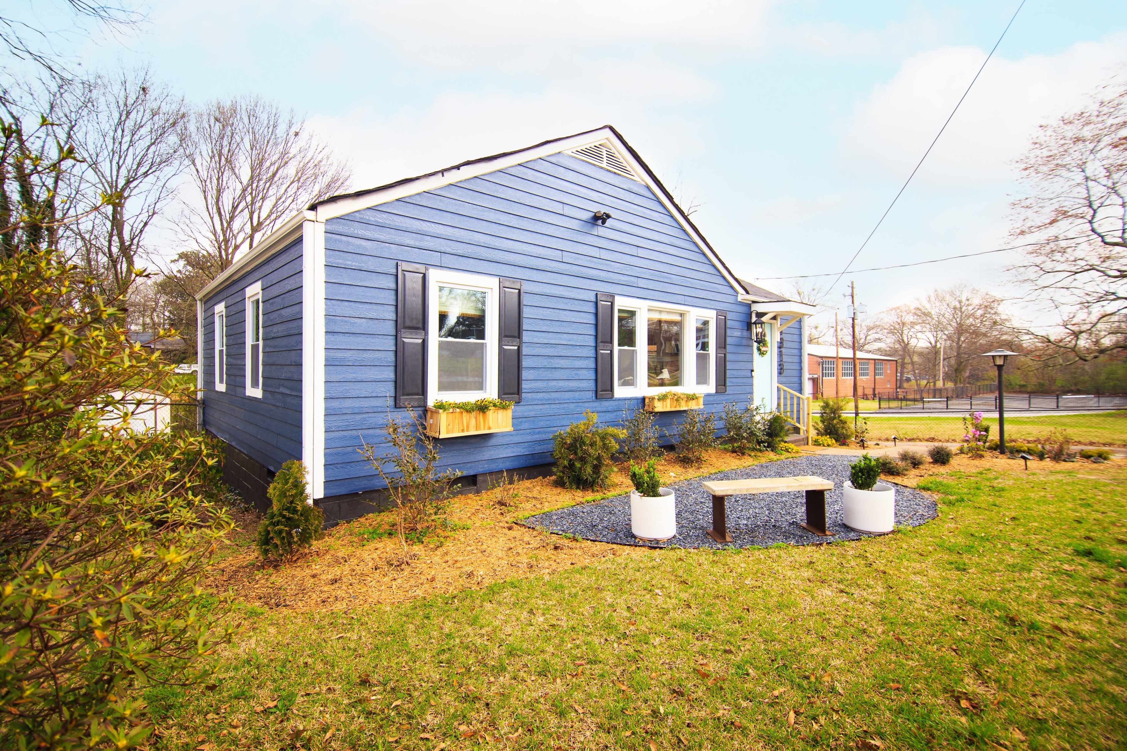 The image shows a blue house with decorative window boxes, surrounded by neatly trimmed landscaping and a small sitting area featuring white planters.