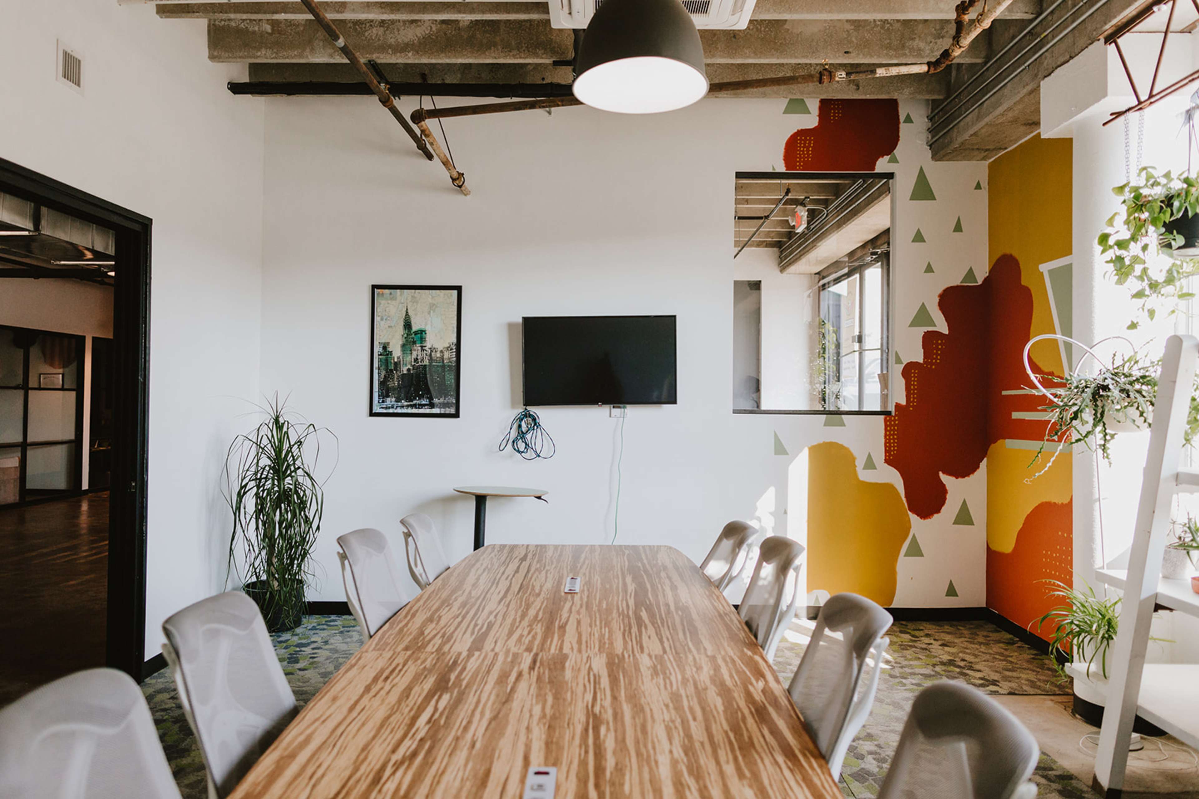 A modern conference room features a long wooden table, a wall-mounted TV, and colorful geometric artwork alongside potted plants.