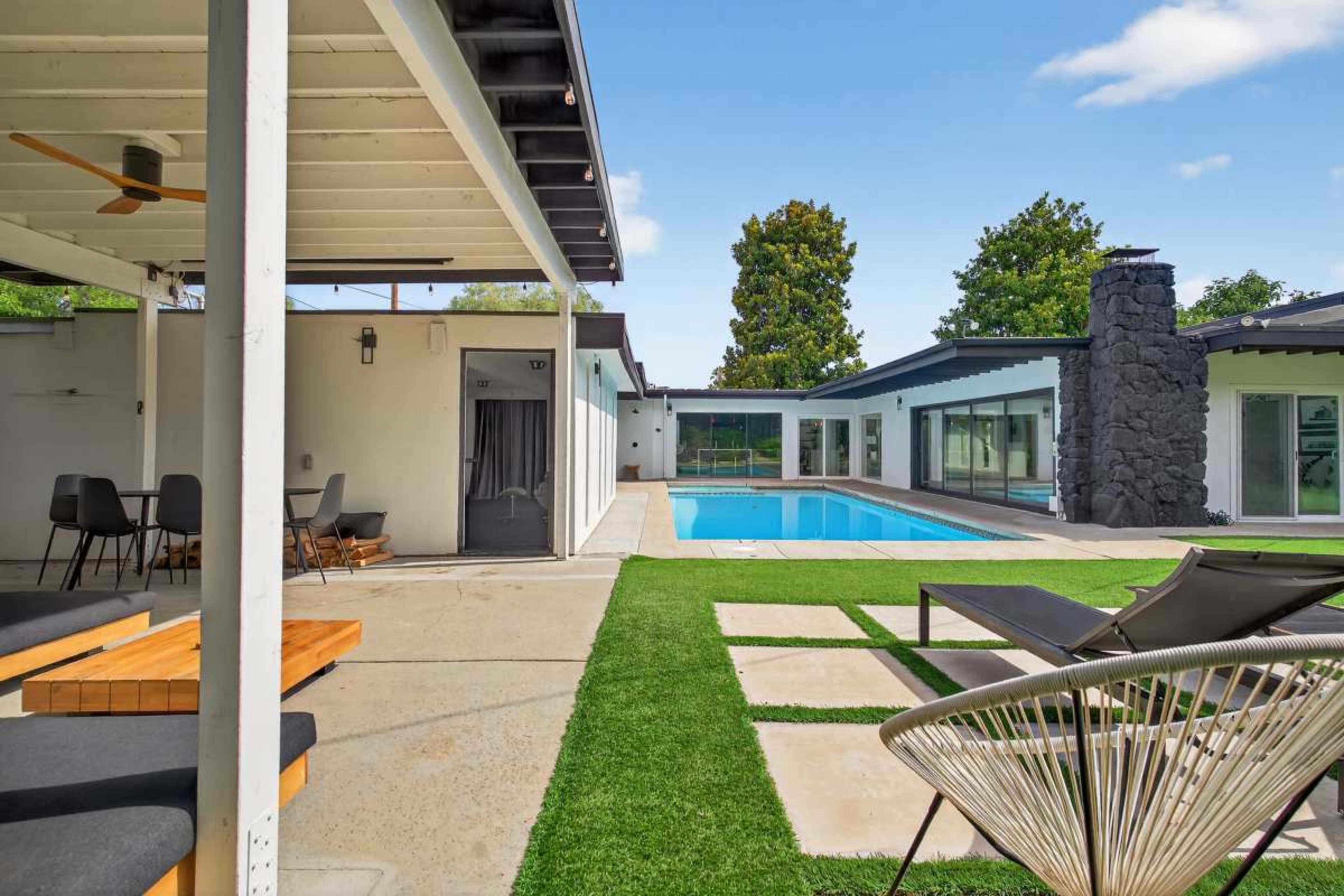The image depicts a modern outdoor living space featuring a pool, lounge chairs, and a stone fireplace, surrounded by greenery and contemporary architecture.