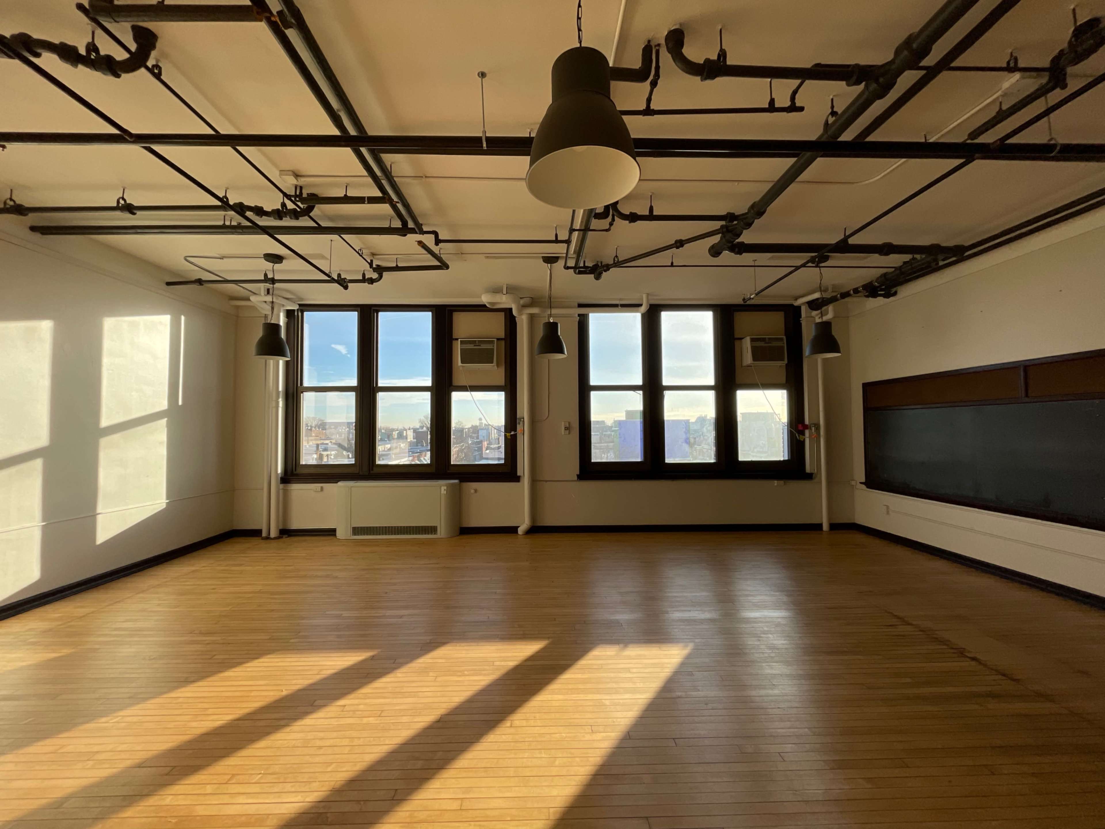 A spacious room with large windows, wooden flooring, and overhead pipes, featuring sunlight casting shadows on the floor.