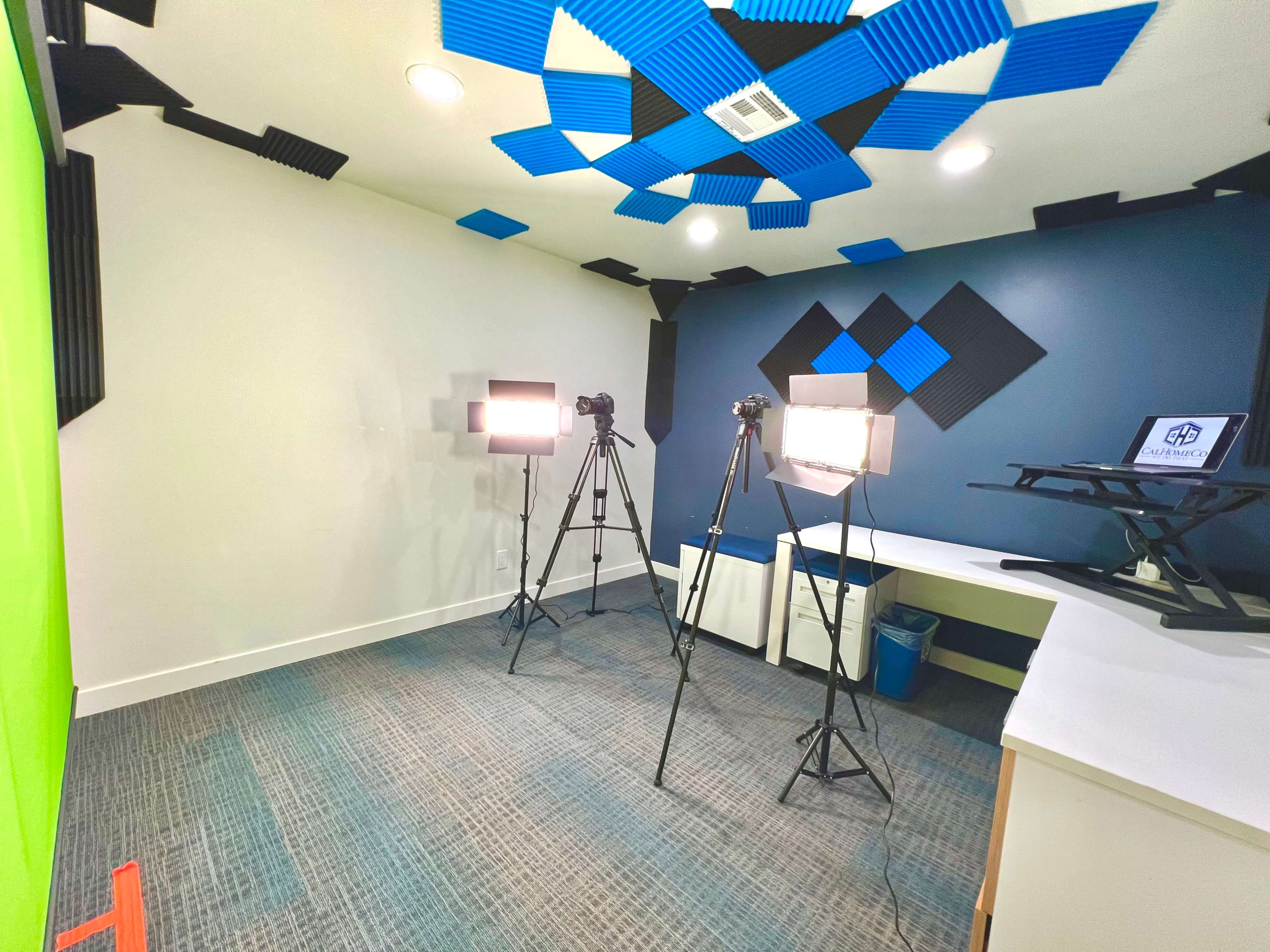 A room equipped for video production, featuring a green screen, two camera tripods with lights, and a desk with a laptop.