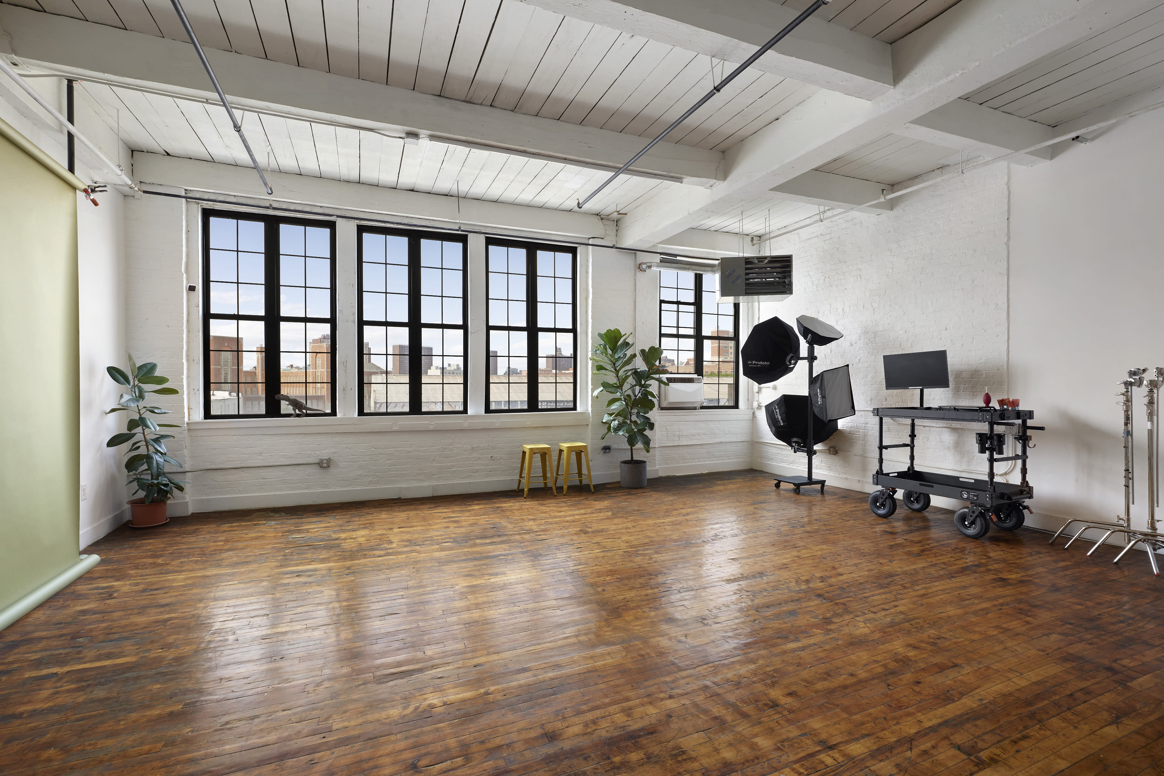 A spacious photography studio with large windows, wooden floors, and various lighting equipment set up in the corner.