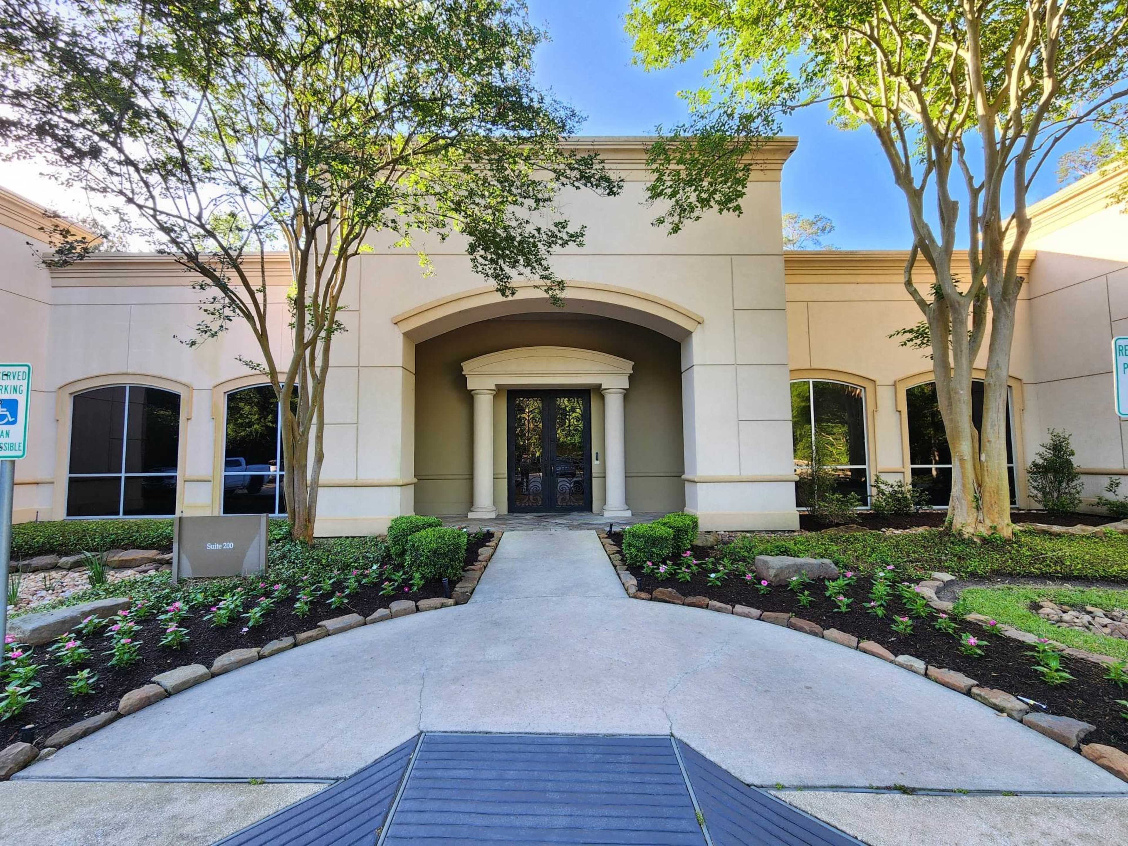 The entrance of a building framed by trees and landscaped bushes along a paved walkway.