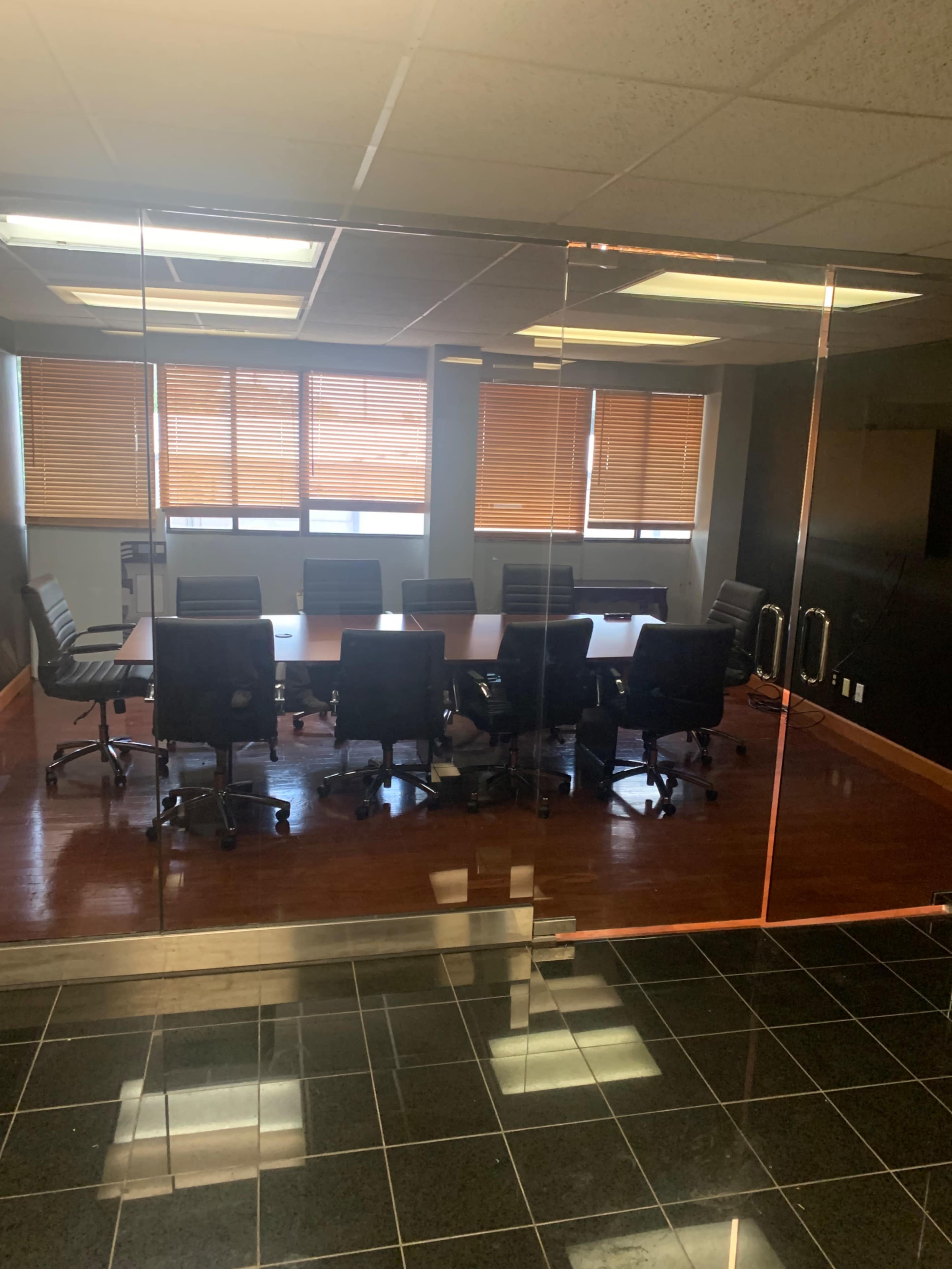 The image shows a conference room with a large rectangular table surrounded by black rolling chairs and large windows with closed blinds.