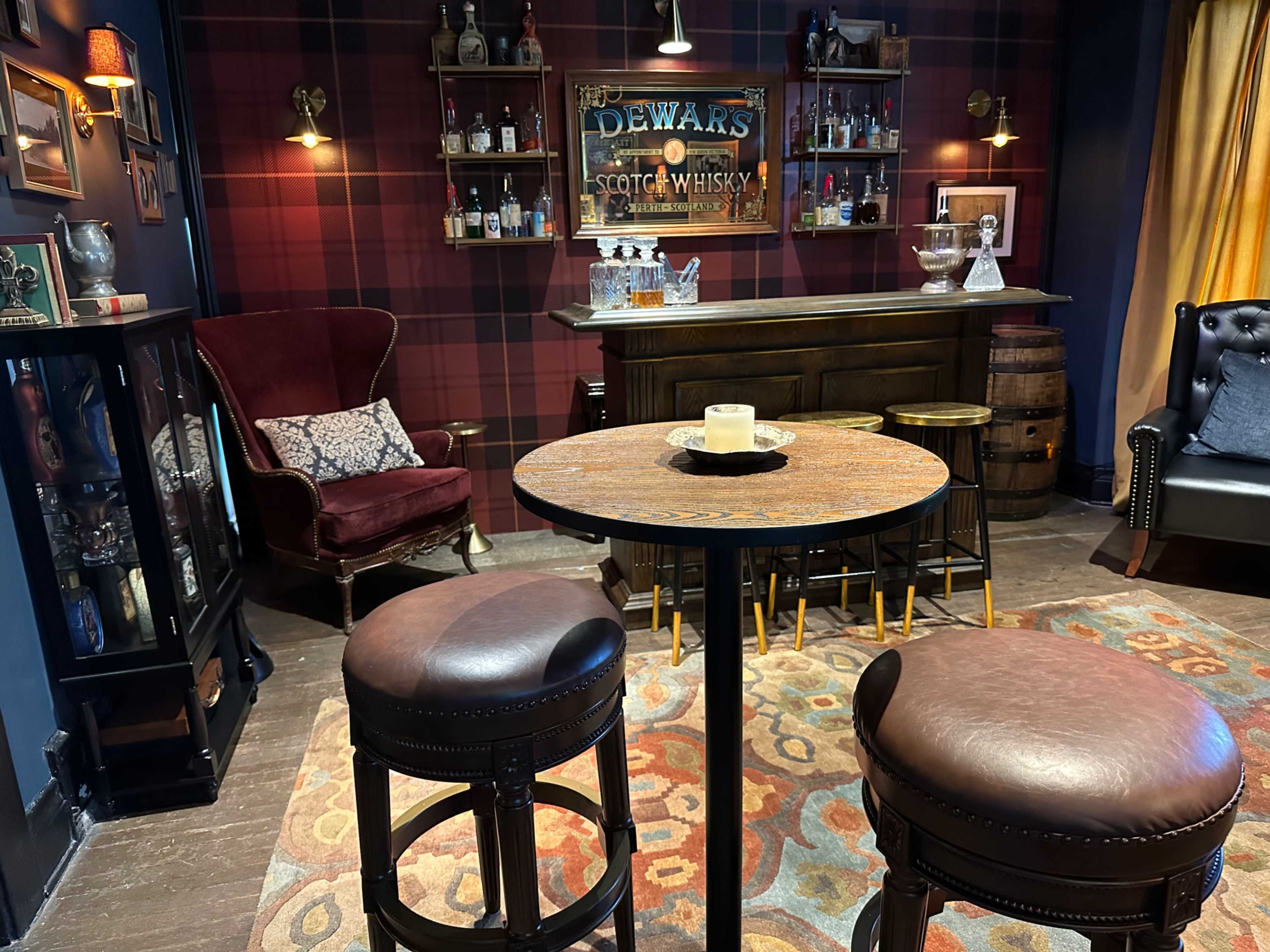 The image shows a cozy bar setup with a dark wood bar, a bottle display, two high stools, and a red plaid wall design.