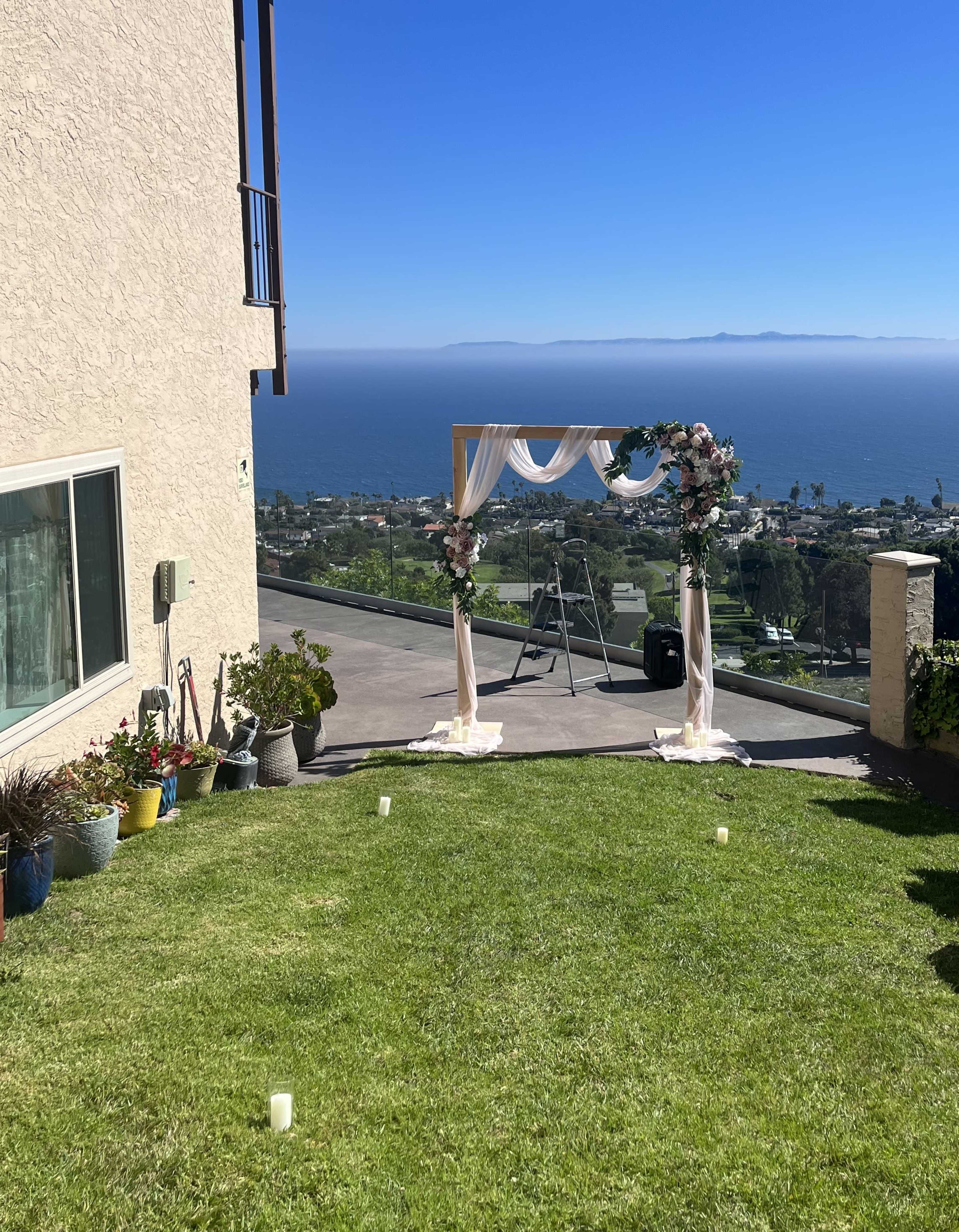 A floral arch is set up on a grassy area overlooking the ocean with distant hills in the background.