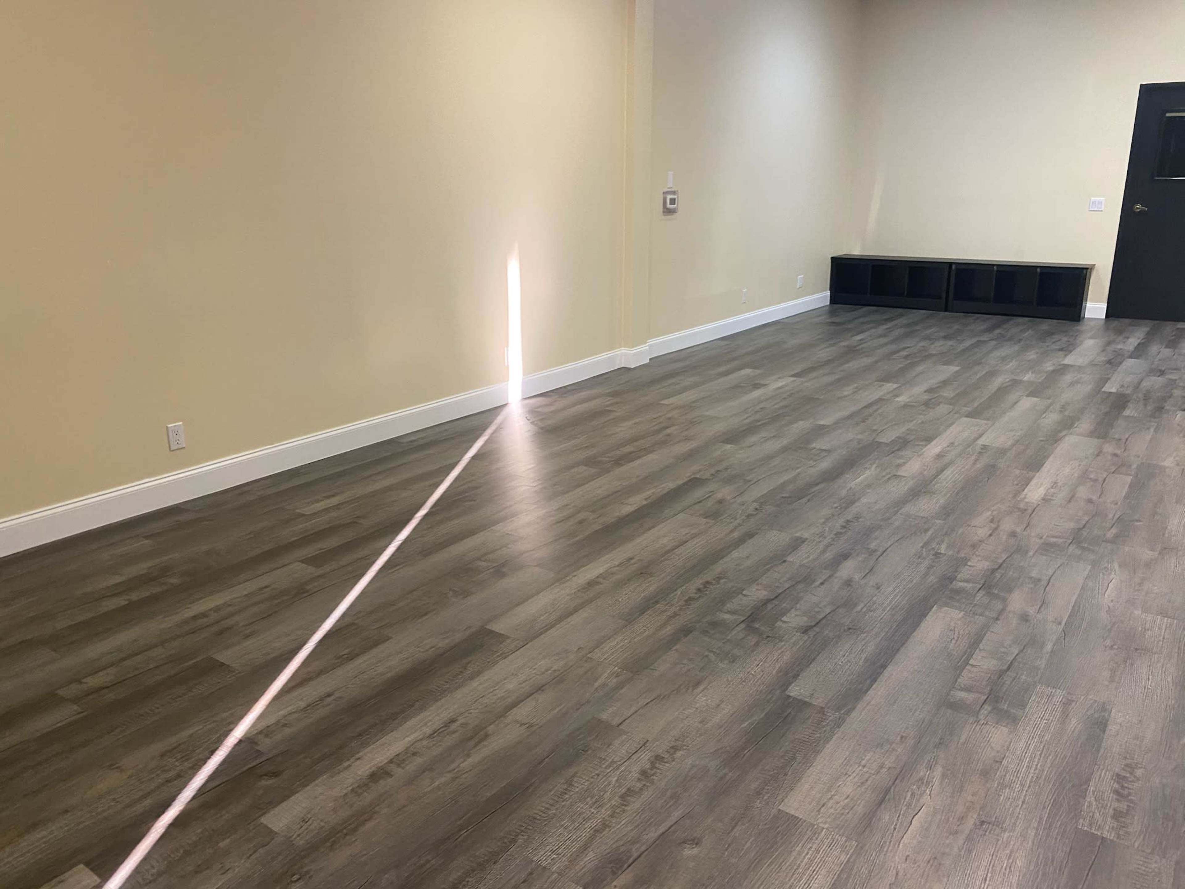 The image shows a spacious room with light-colored walls and a wooden floor, featuring a narrow beam of light coming from a door on one side.