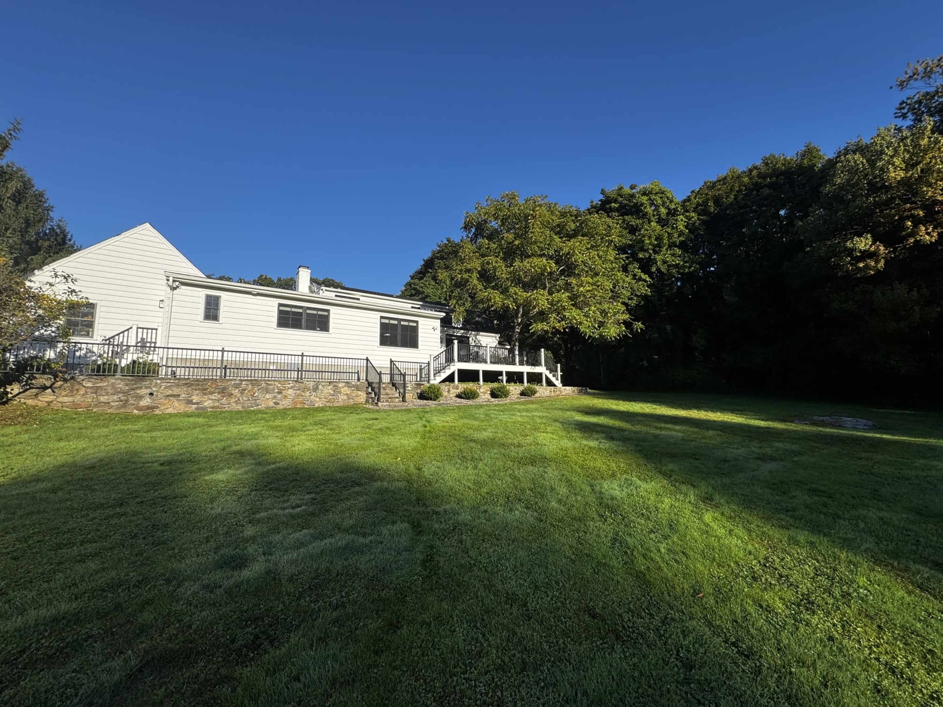A modern house with a stone foundation is situated on a grassy lawn, surrounded by trees under a clear blue sky.