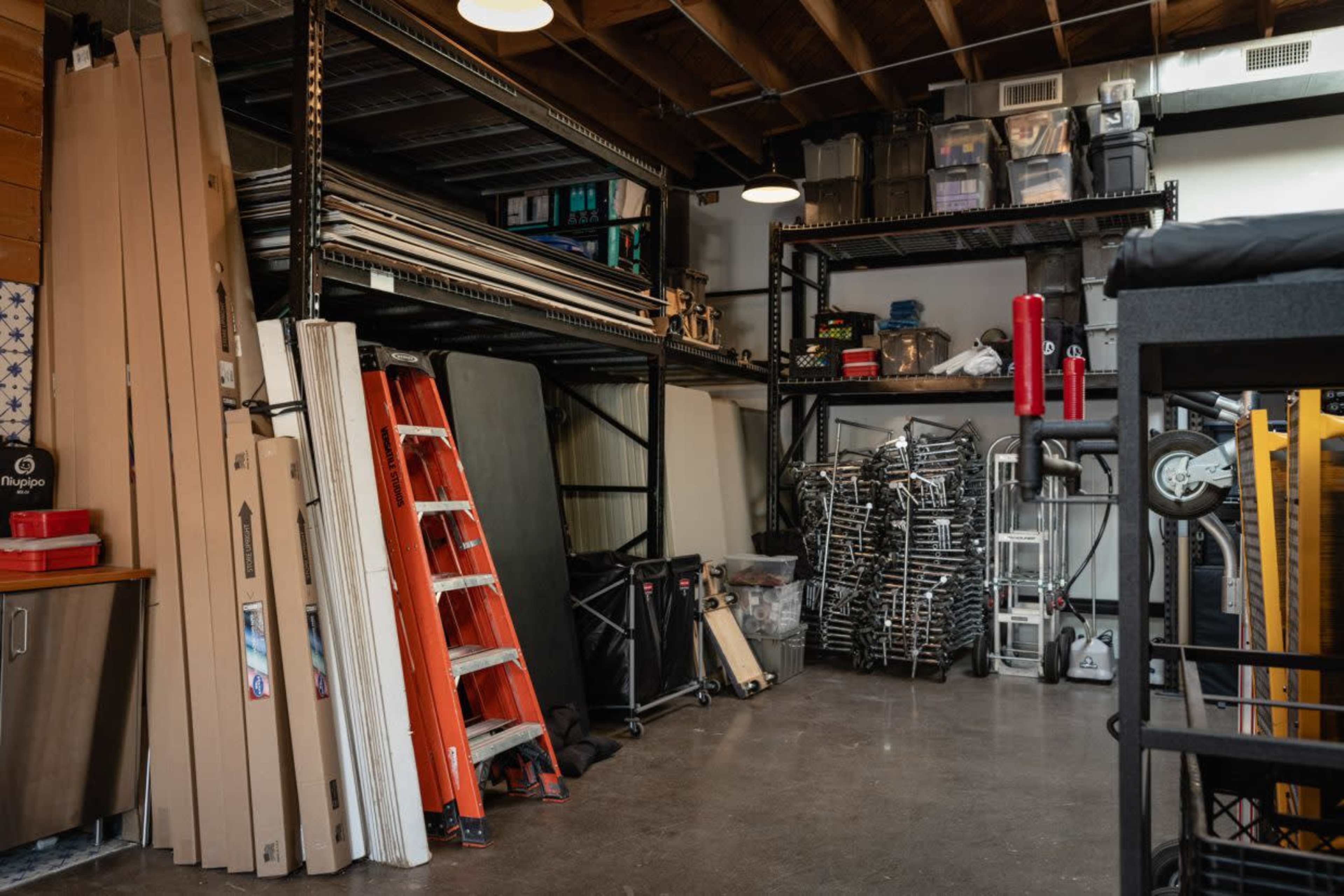 The image shows a storage area filled with various materials including ladders, stacking chairs, shelving units, and plastic containers.