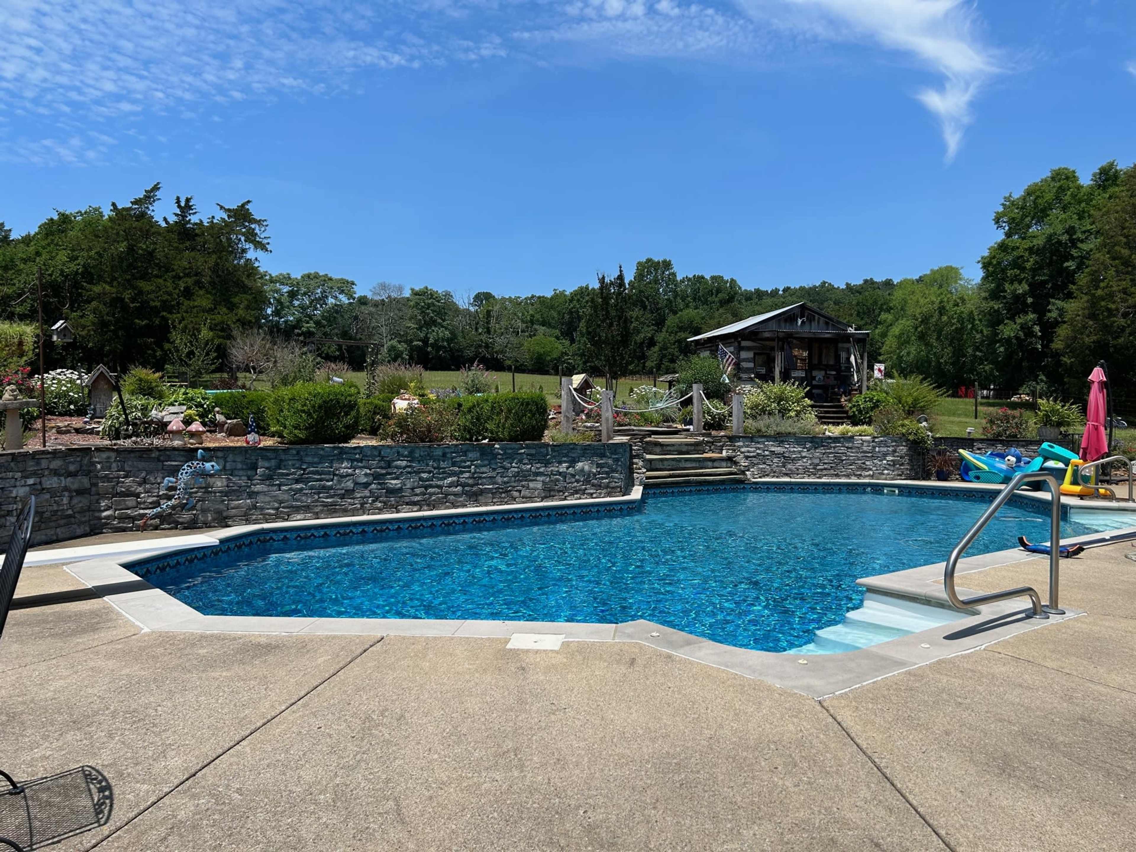 The image shows a clear blue swimming pool surrounded by a concrete deck, with a stone wall and a grassy area in the background.