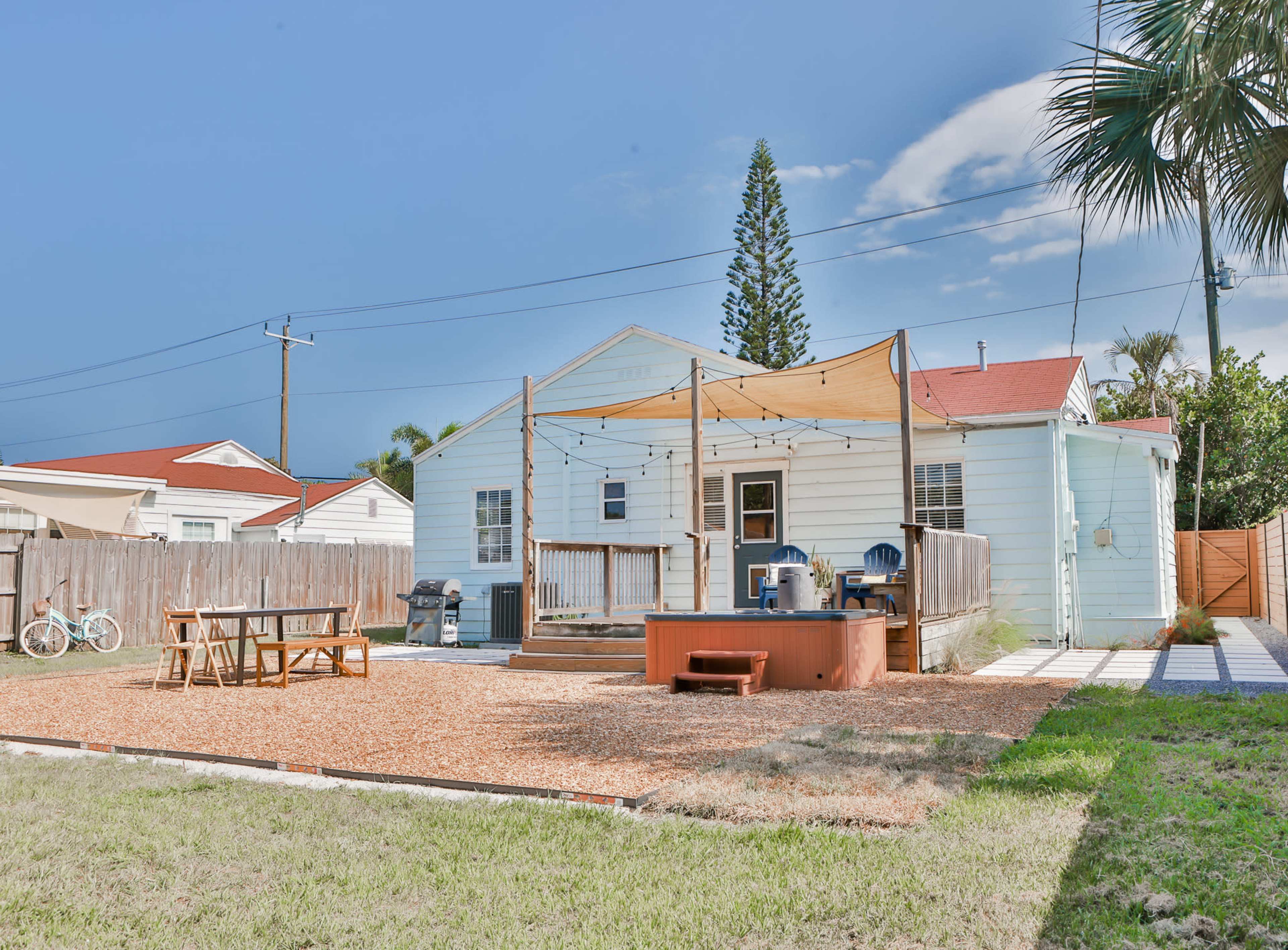 A backyard features a wooden deck with a pergola, a hot tub, and a picnic table surrounded by gravel and grass, with a house and neighboring structures in the background.