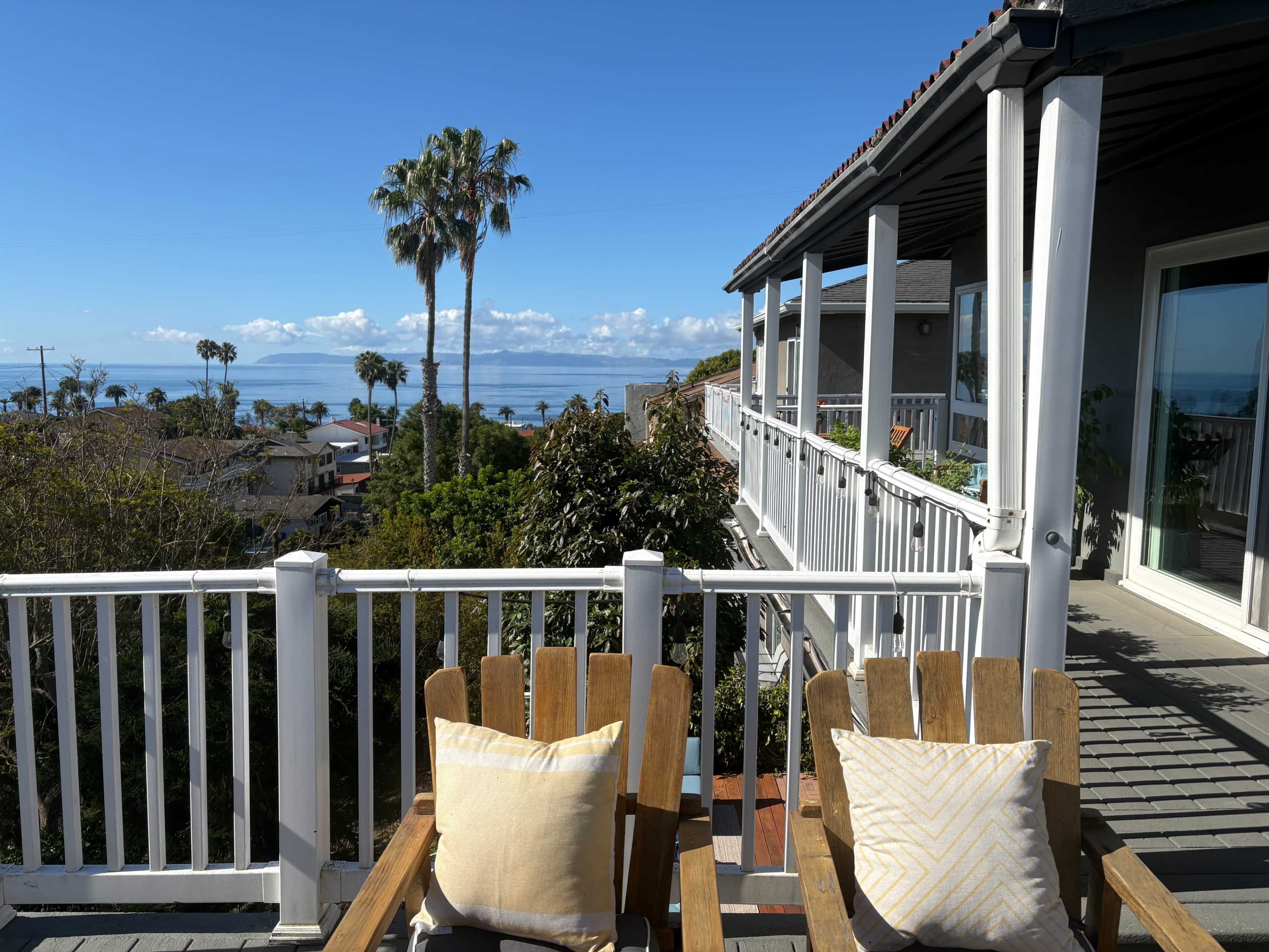 Two wooden chairs with cushions are positioned on a balcony overlooking a coastal view with palm trees and the ocean in the distance.