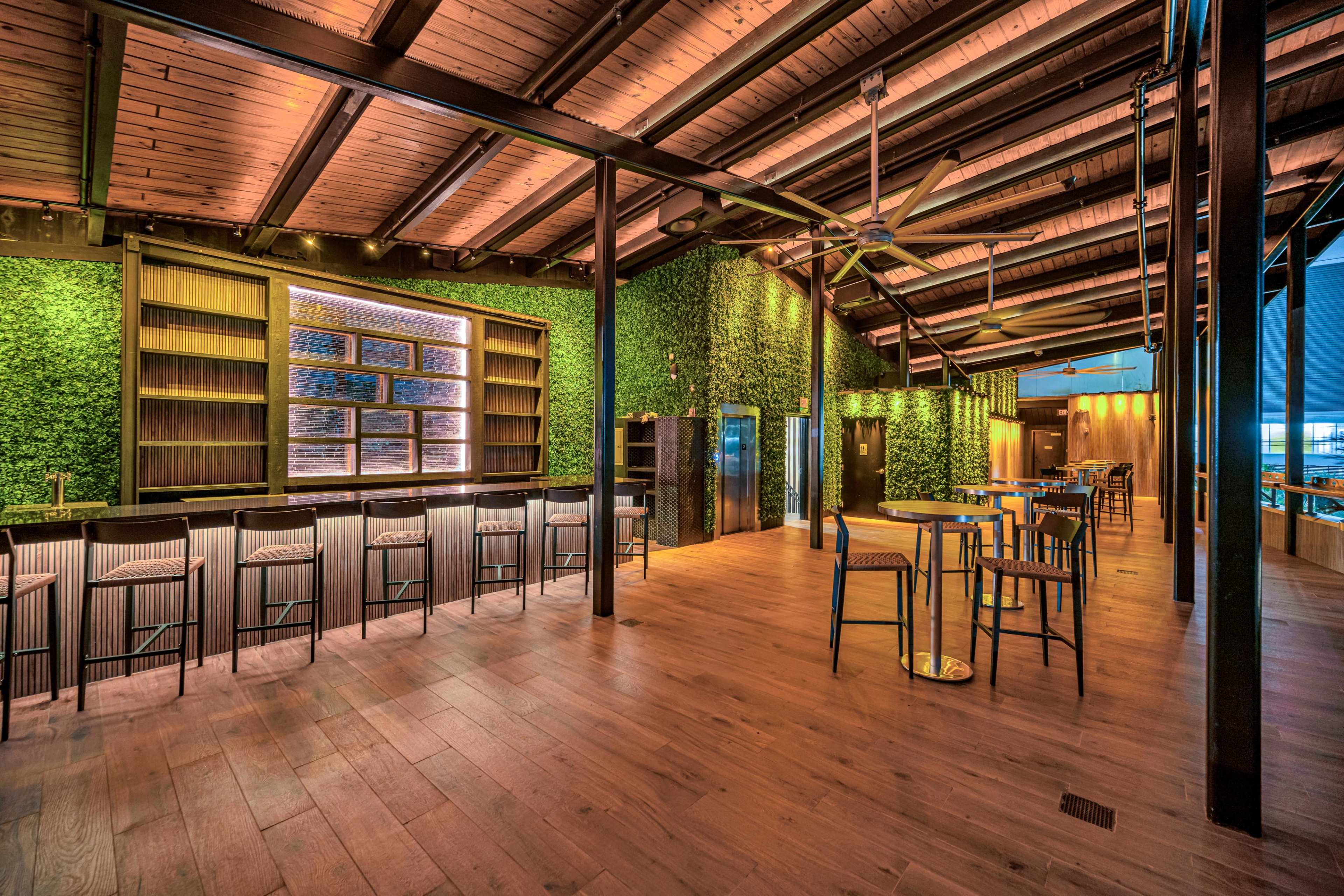 The image shows a modern bar interior with green wall plant decorations, wooden floors, and a row of high chairs along a sleek counter.