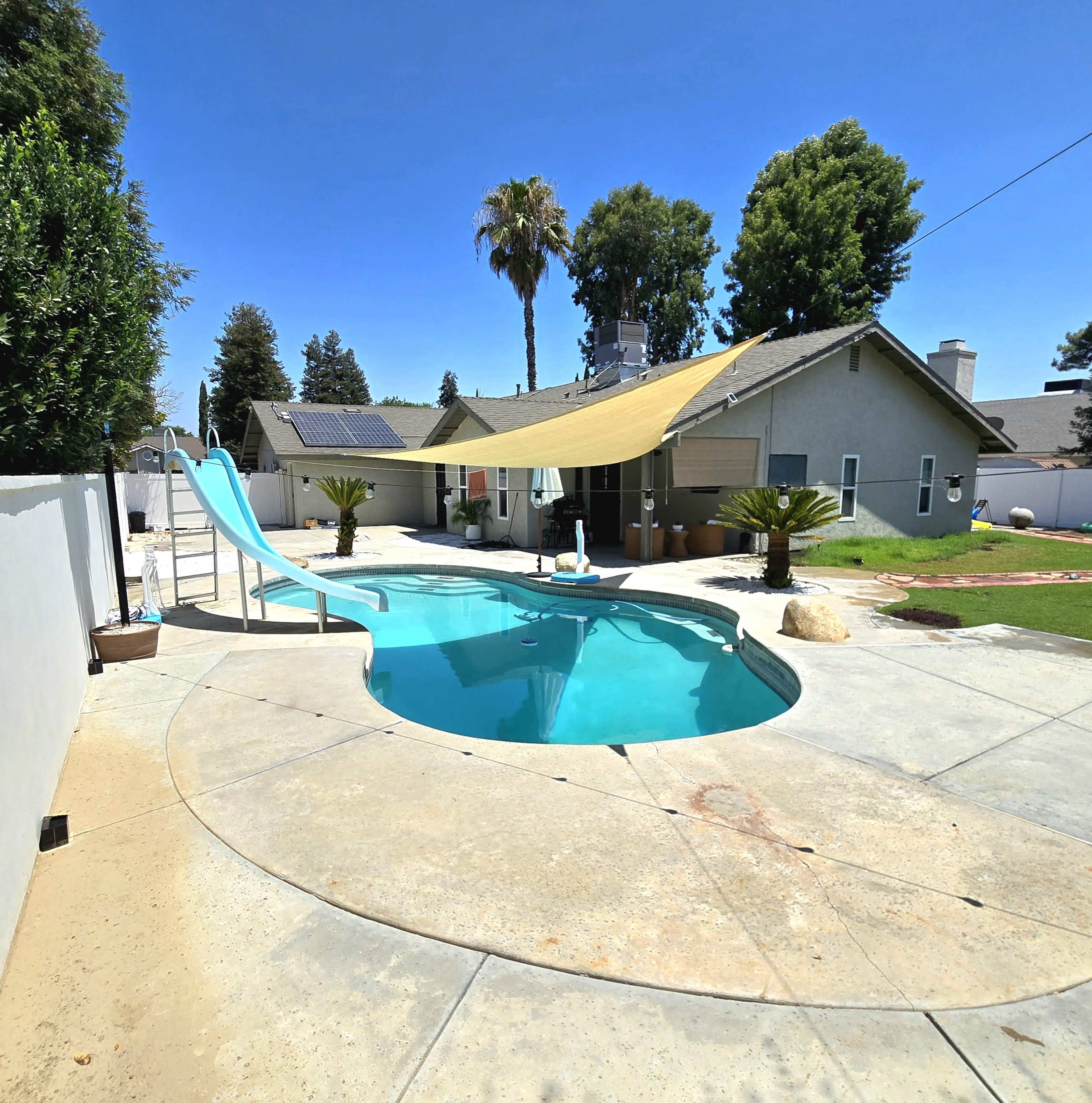 A backyard swimming pool with a slide, surrounded by a landscaped garden, and a shaded area overhead.