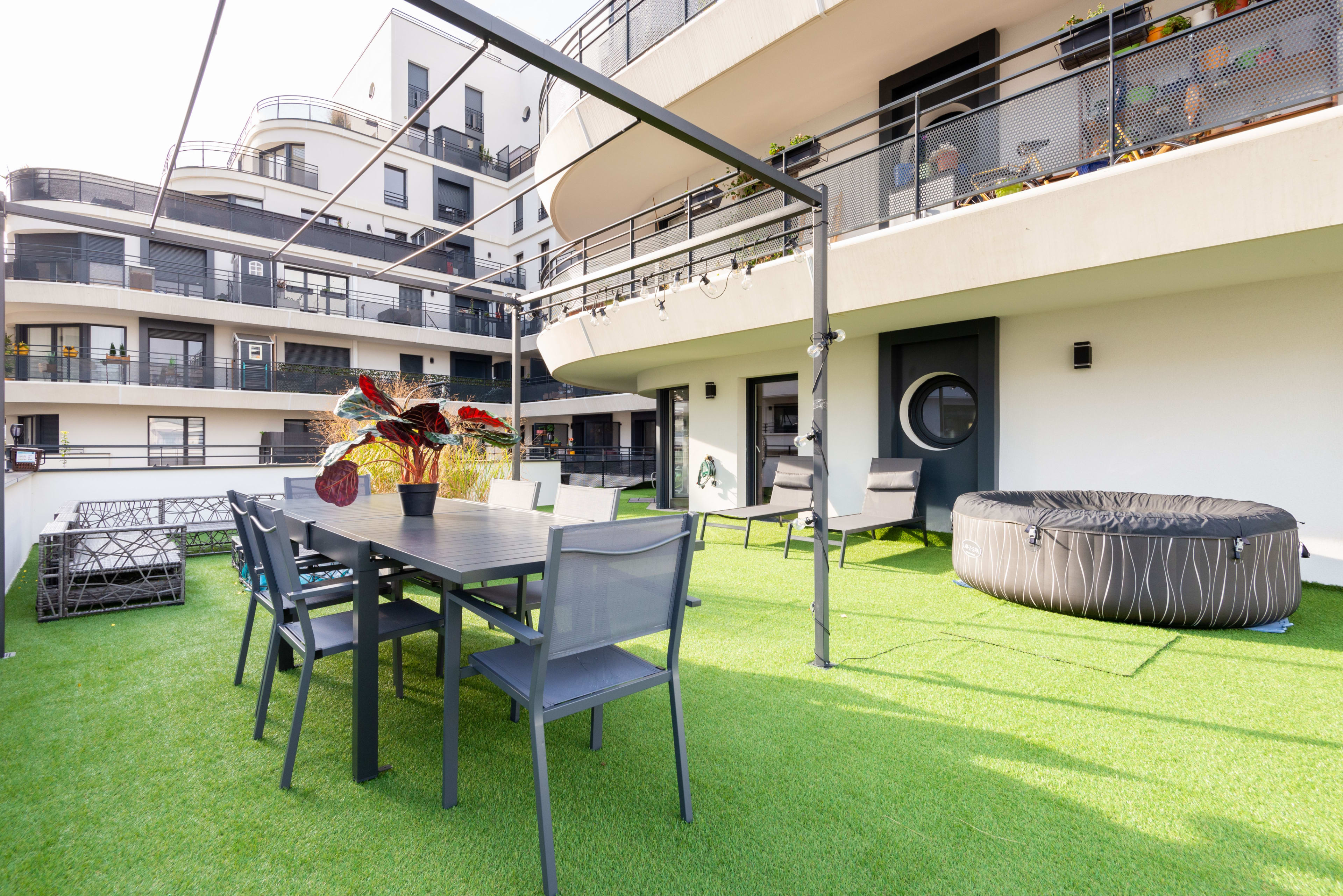 A modern outdoor terrace features a dining table with chairs, lounge seating, and a circular hot tub, surrounded by sleek apartment balconies.