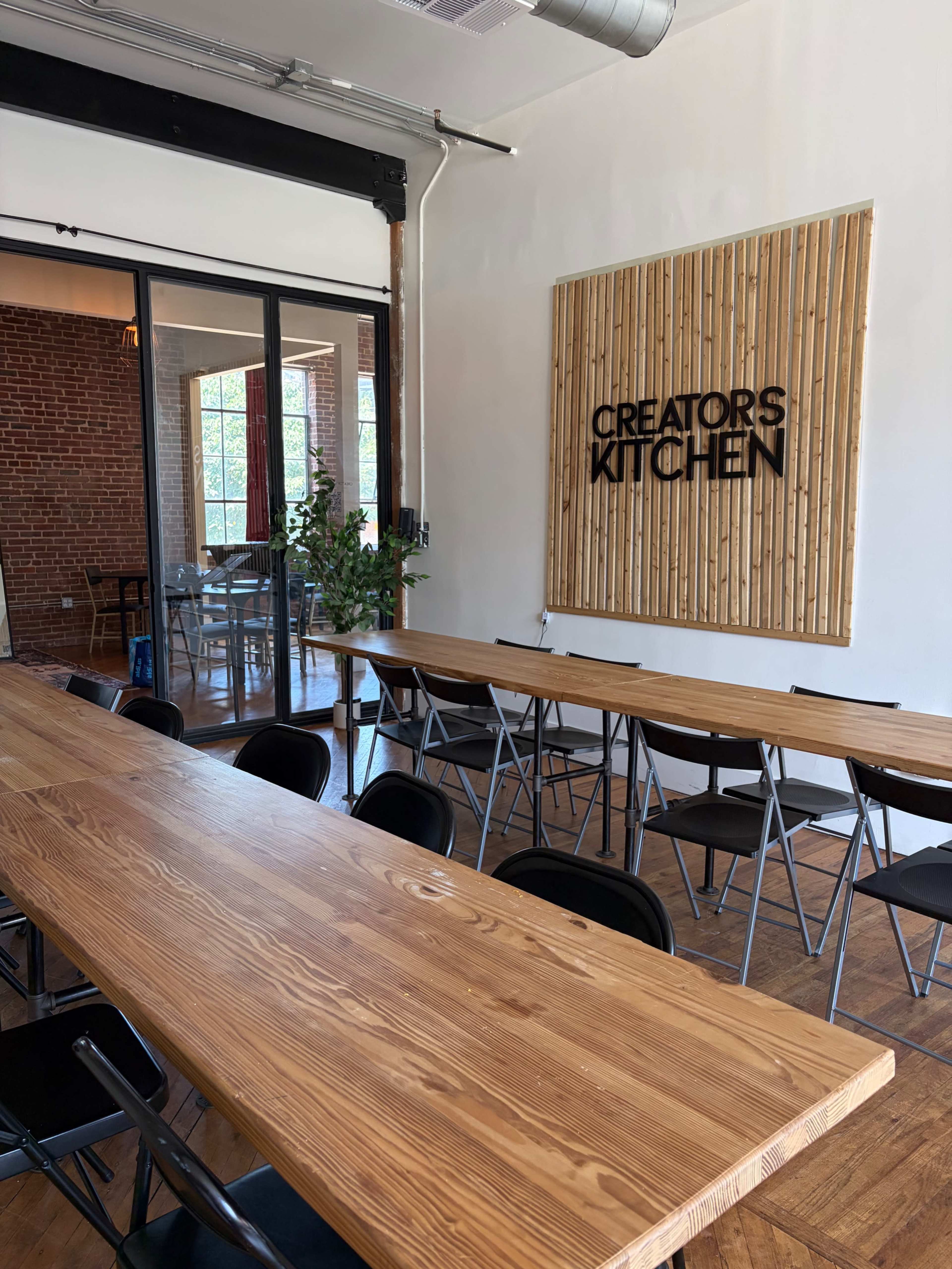 The image shows a spacious room with a long wooden table and folding chairs, featuring a wall sign that reads "CREATORS KITCHEN" and exposed brick walls in the background.