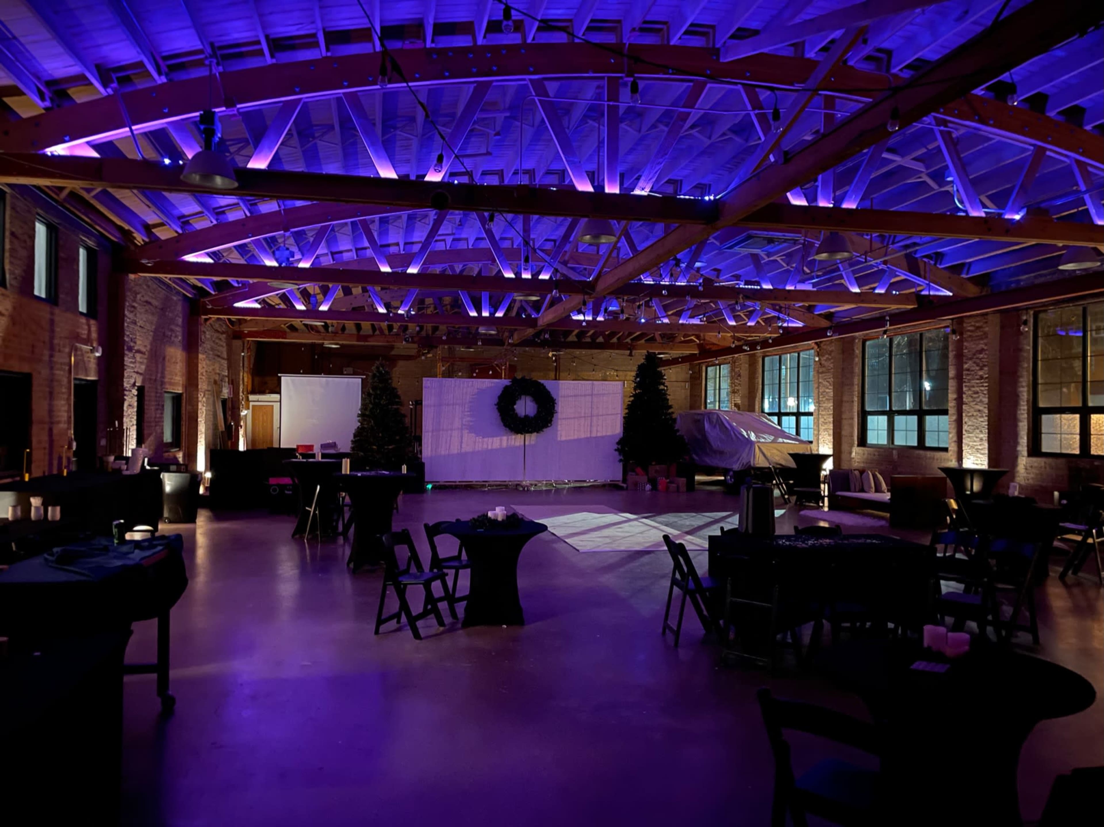 The image shows an indoor event space with a high ceiling, decorated with purple lighting, tables, and Christmas trees.
