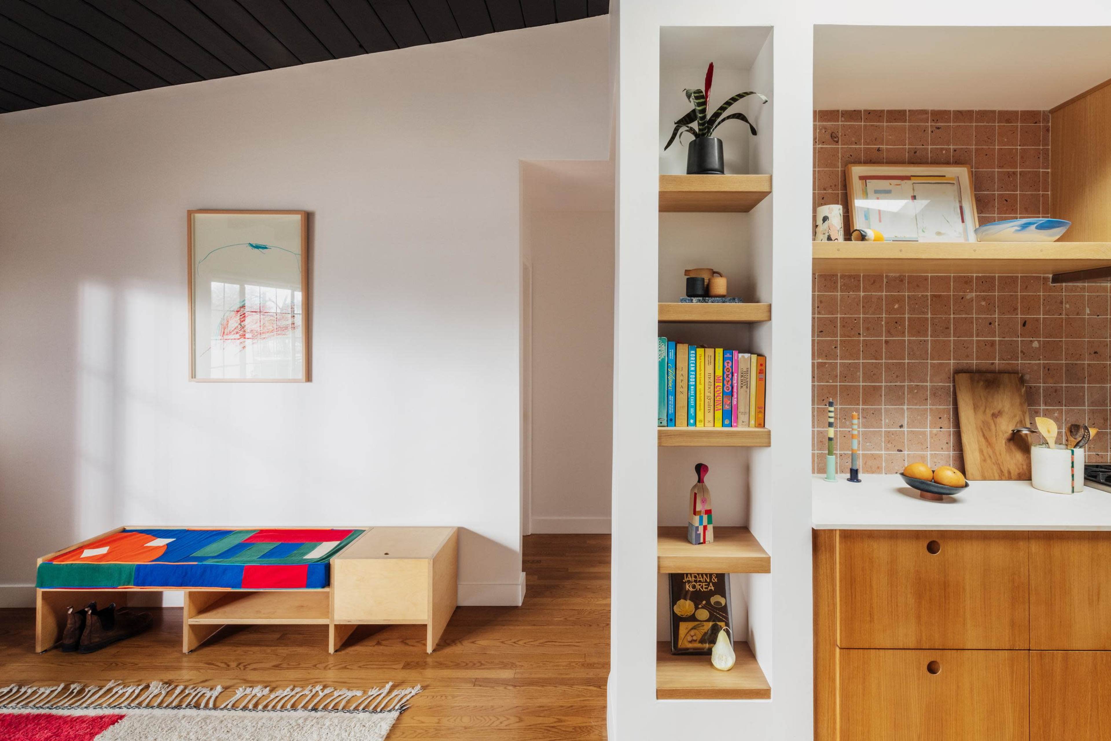 The image shows a modern living space featuring a colorful bench, a shelving unit with books and decor, and a kitchen area with a light wood finish and a tile backsplash.