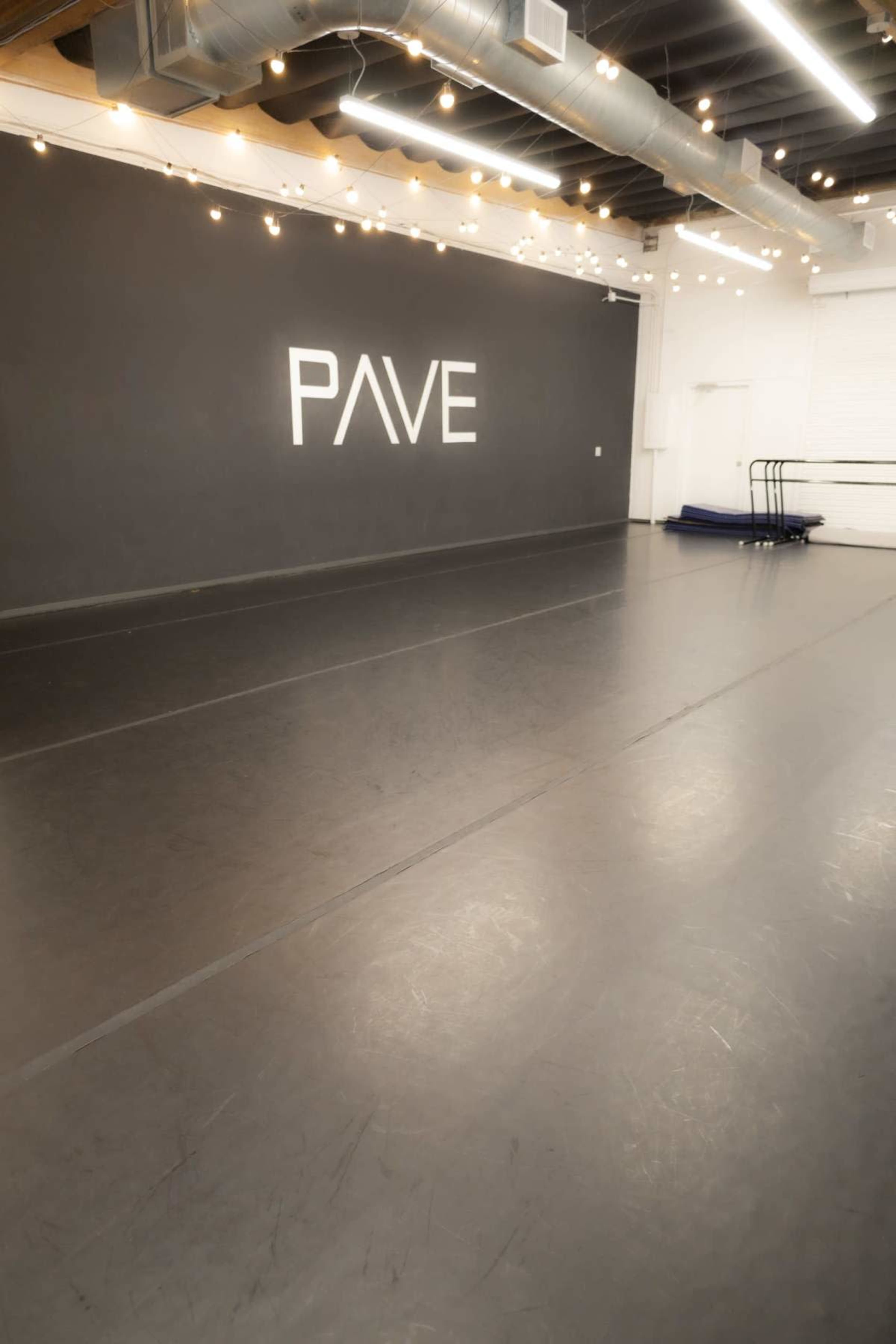 The image shows a dance studio with a dark floor, a wall marked with the word "PAVE," and overhead lights creating a warm ambiance.