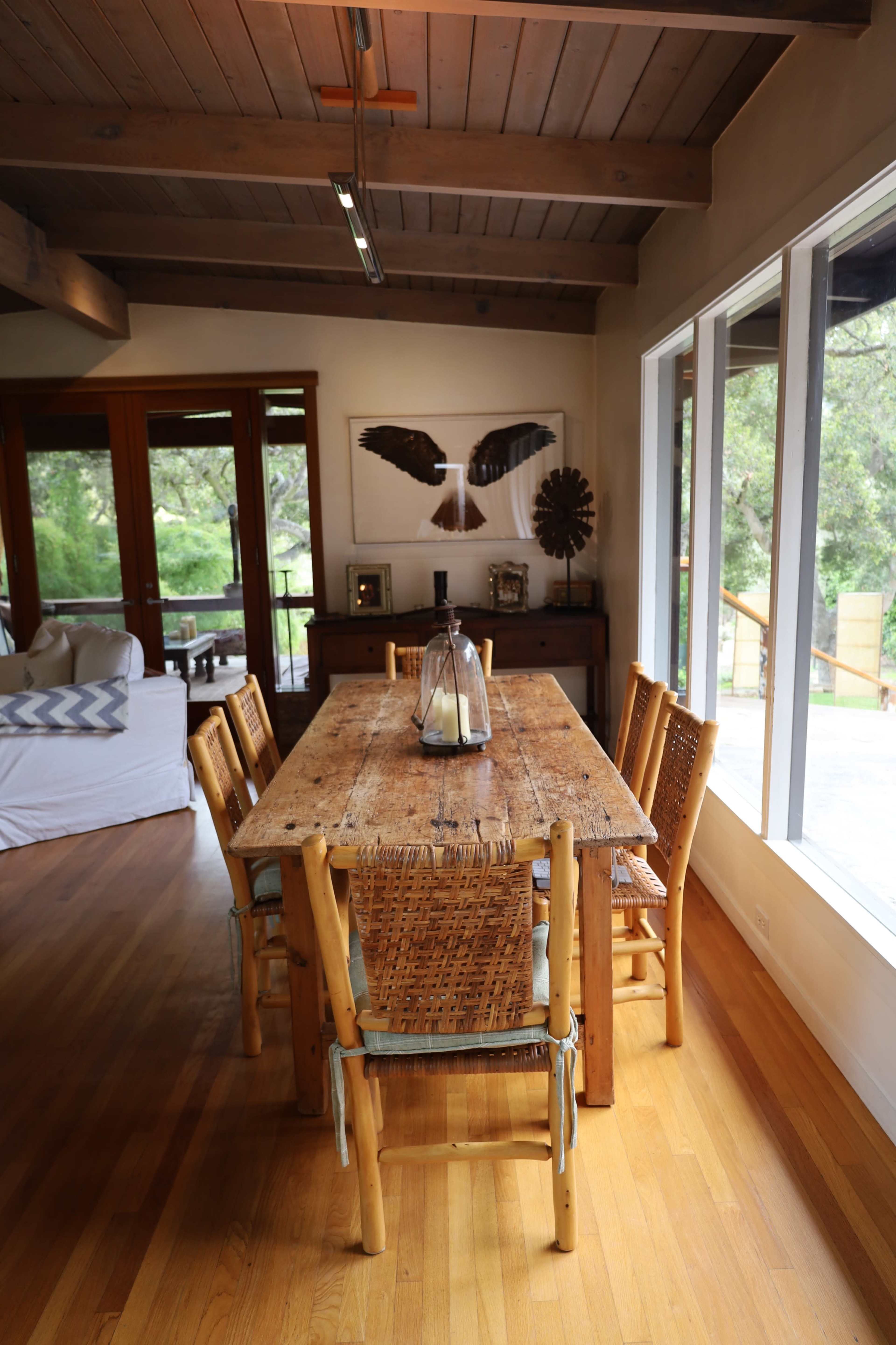A wooden dining table with six chairs is set in a well-lit room featuring large windows and rustic decor.