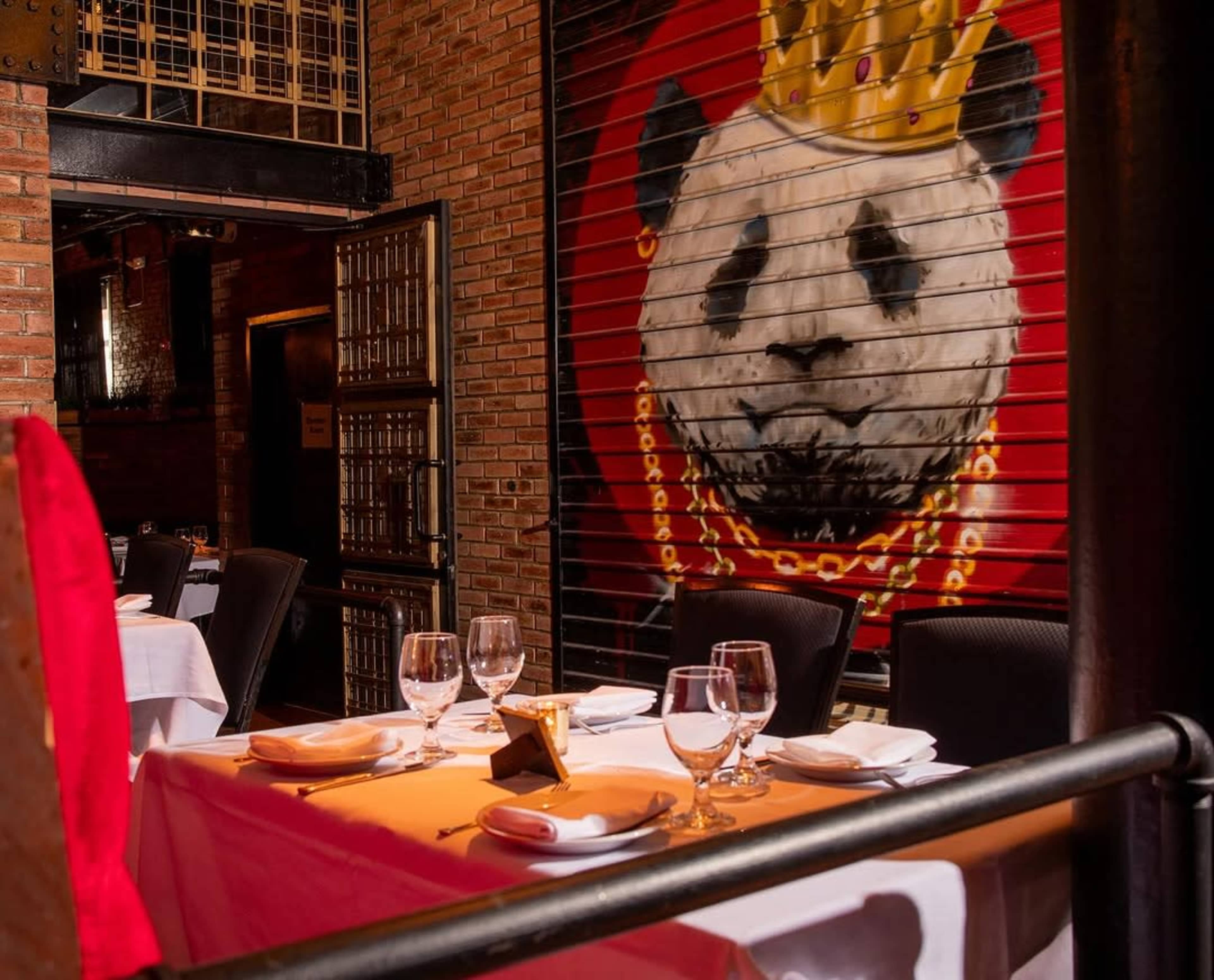 A dining area is set with white tablecloths and elegant glassware in front of a large mural of a panda wearing a crown and jewelry on a brick wall.
