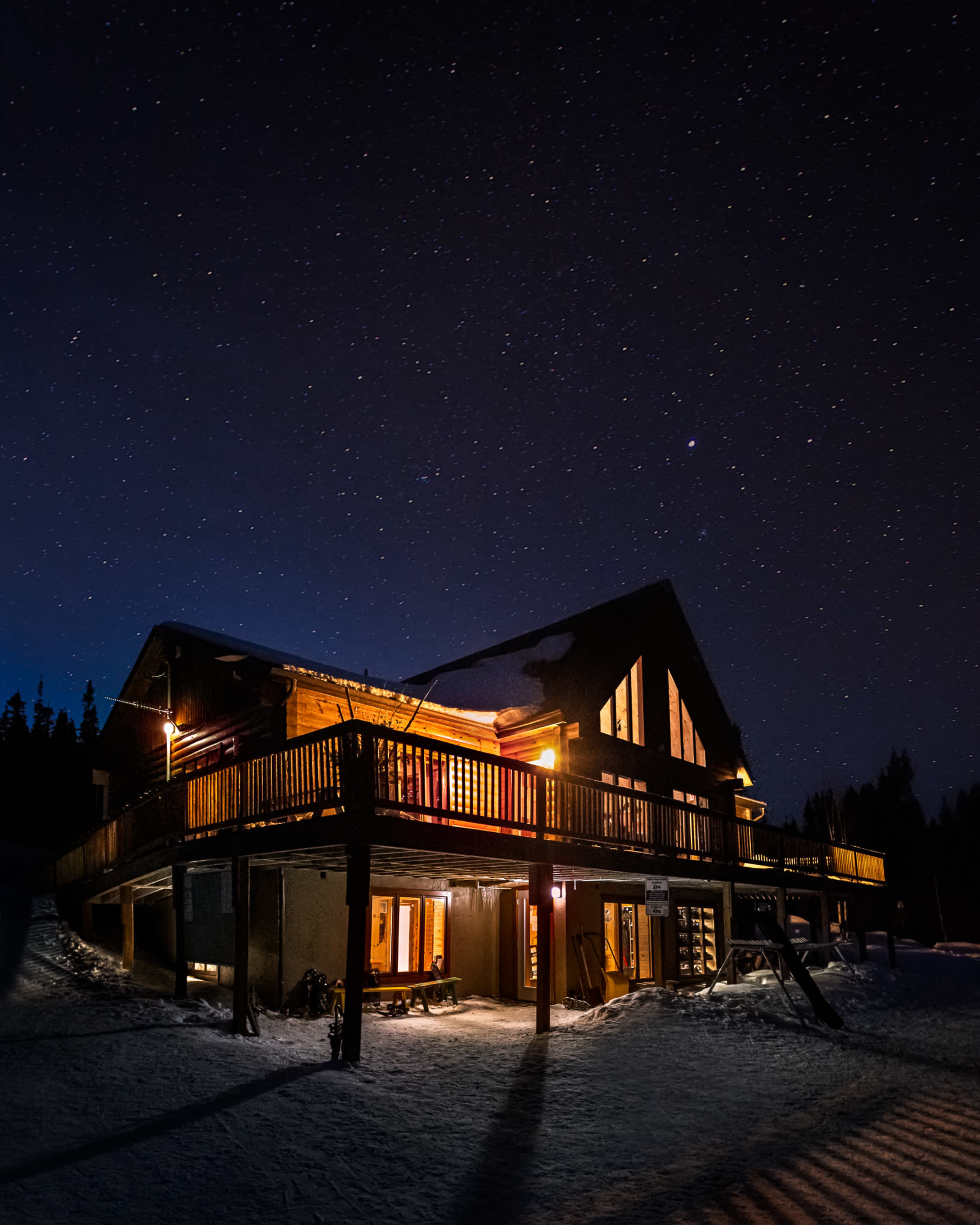 A wooden chalet with illuminated windows stands in a snowy landscape under a starry night sky.