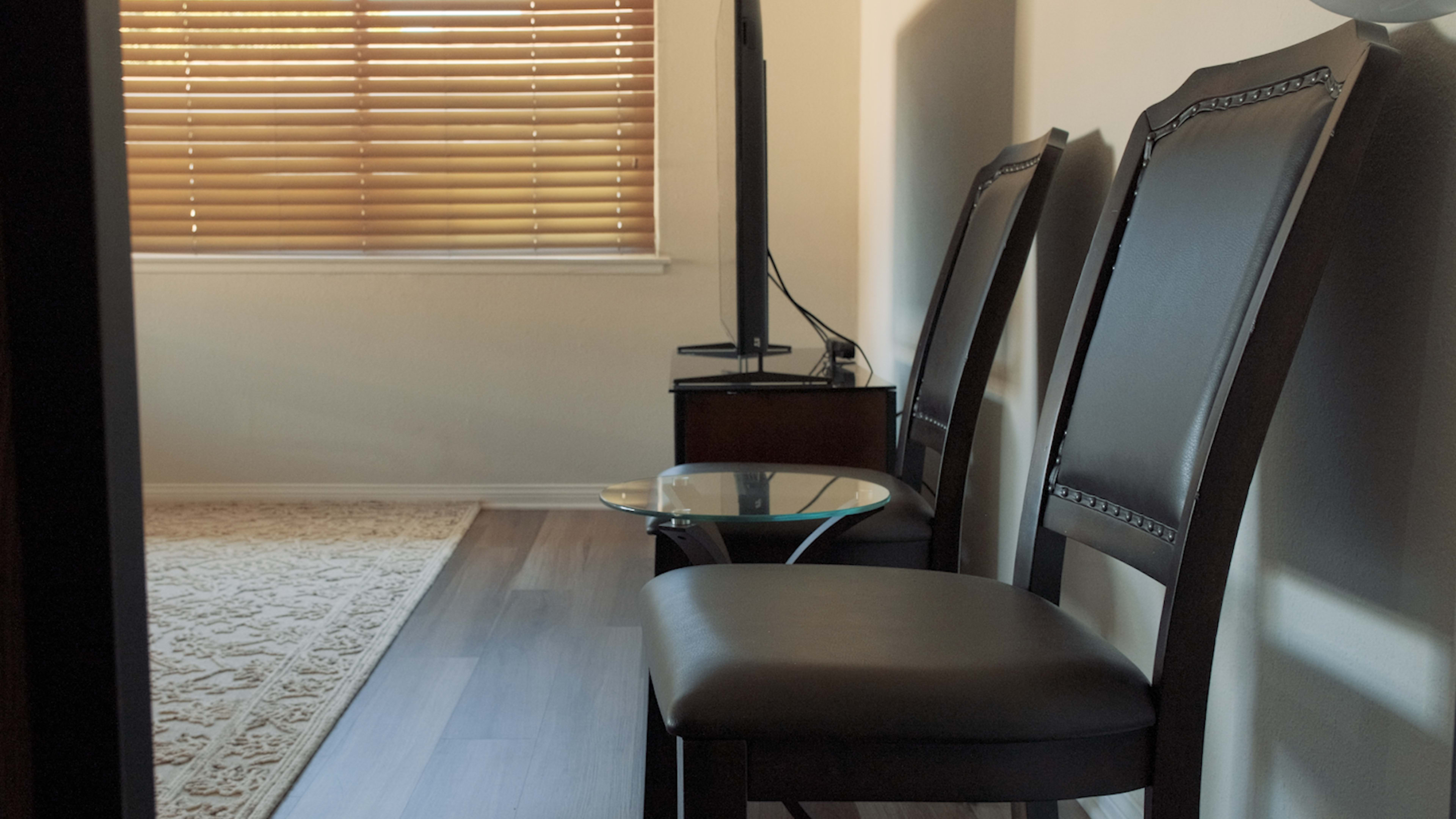 Two black chairs next to a small glass table in a room with wooden flooring and a window covered by blinds.