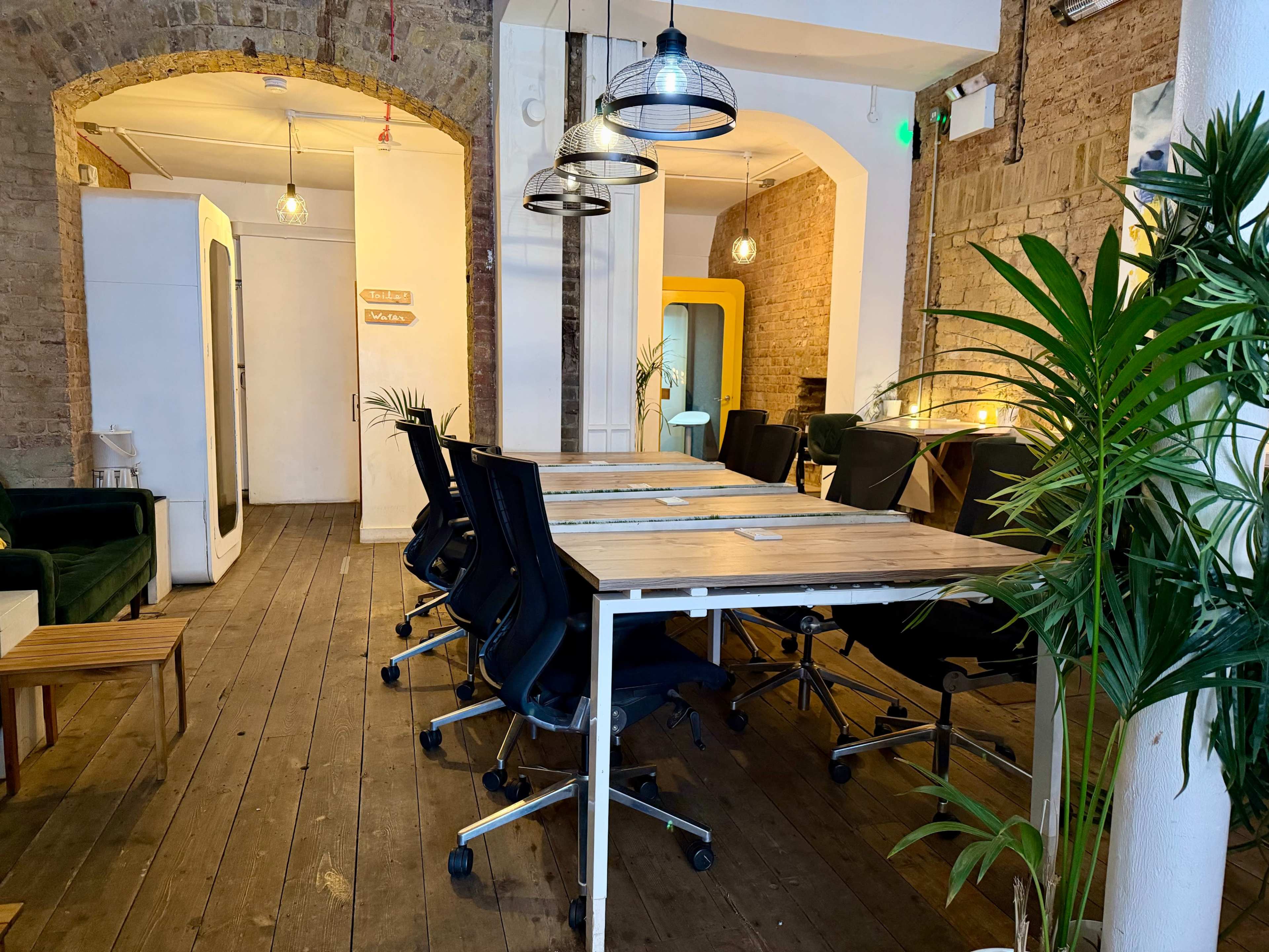 The image shows a spacious, modern workspace with a long table surrounded by black office chairs, brick walls, and decorative plants.