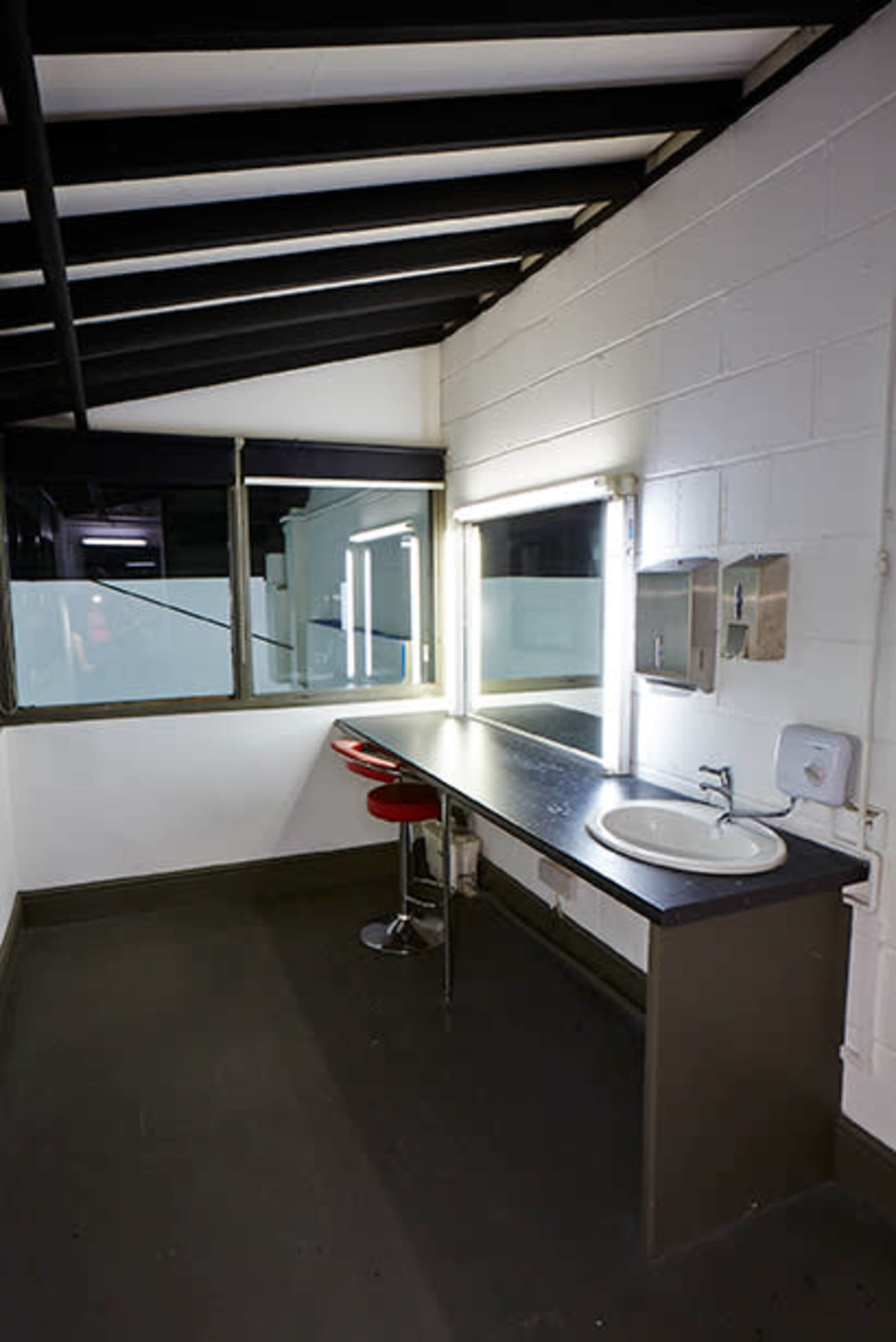 A minimalist restroom with a sink, a red stool, and a large mirror illuminated by fluorescent lights.