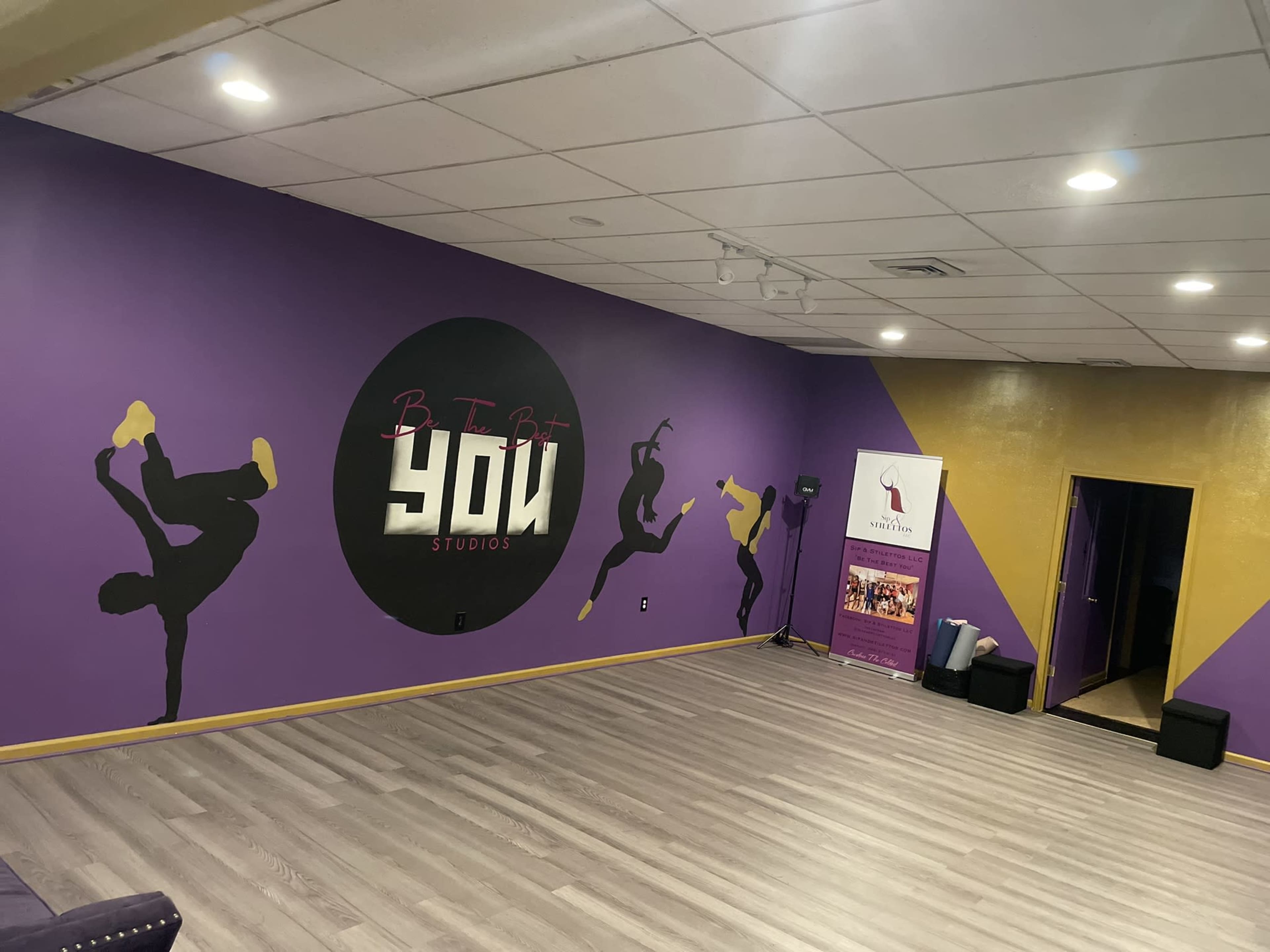 The image shows a dance studio with a purple wall featuring a circular logo that reads "Be The Bit YOU Studios," and a yellow accent wall with an entrance to another room.