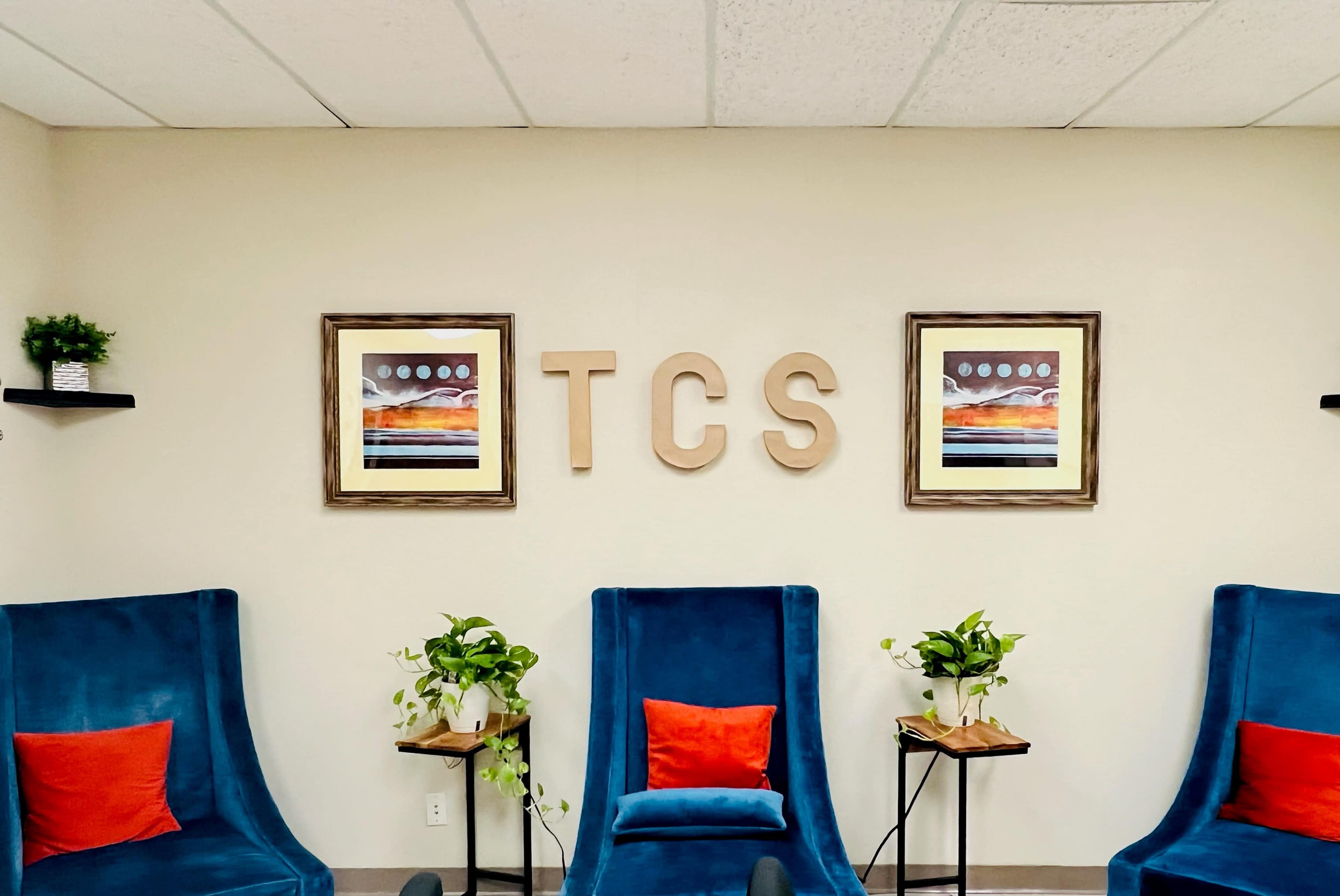 The image shows a waiting area with three blue chairs, two framed artworks on the wall, and wooden letters spelling "TCS" above them.