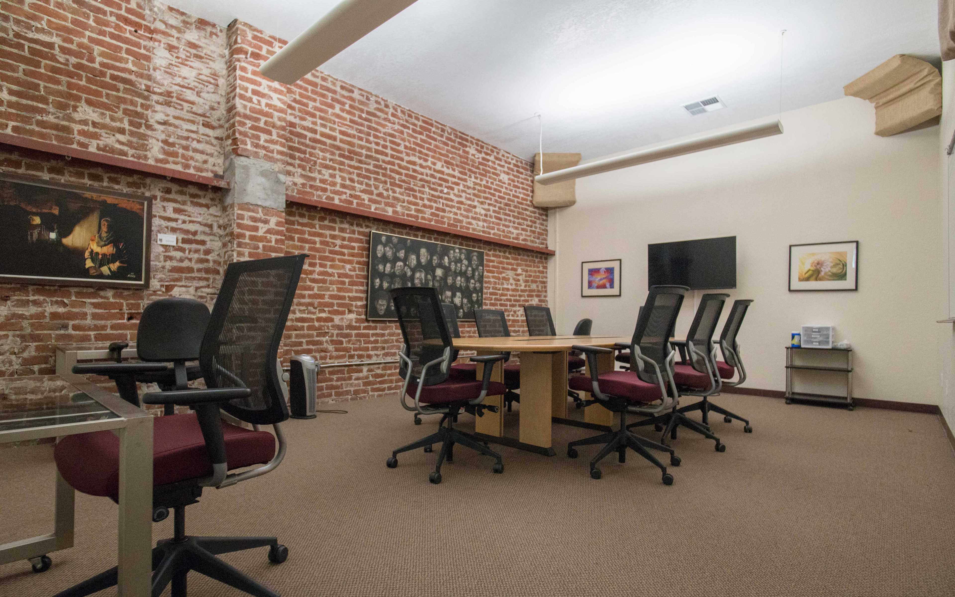 A conference room features a wooden table surrounded by office chairs, with brick walls and framed artwork.