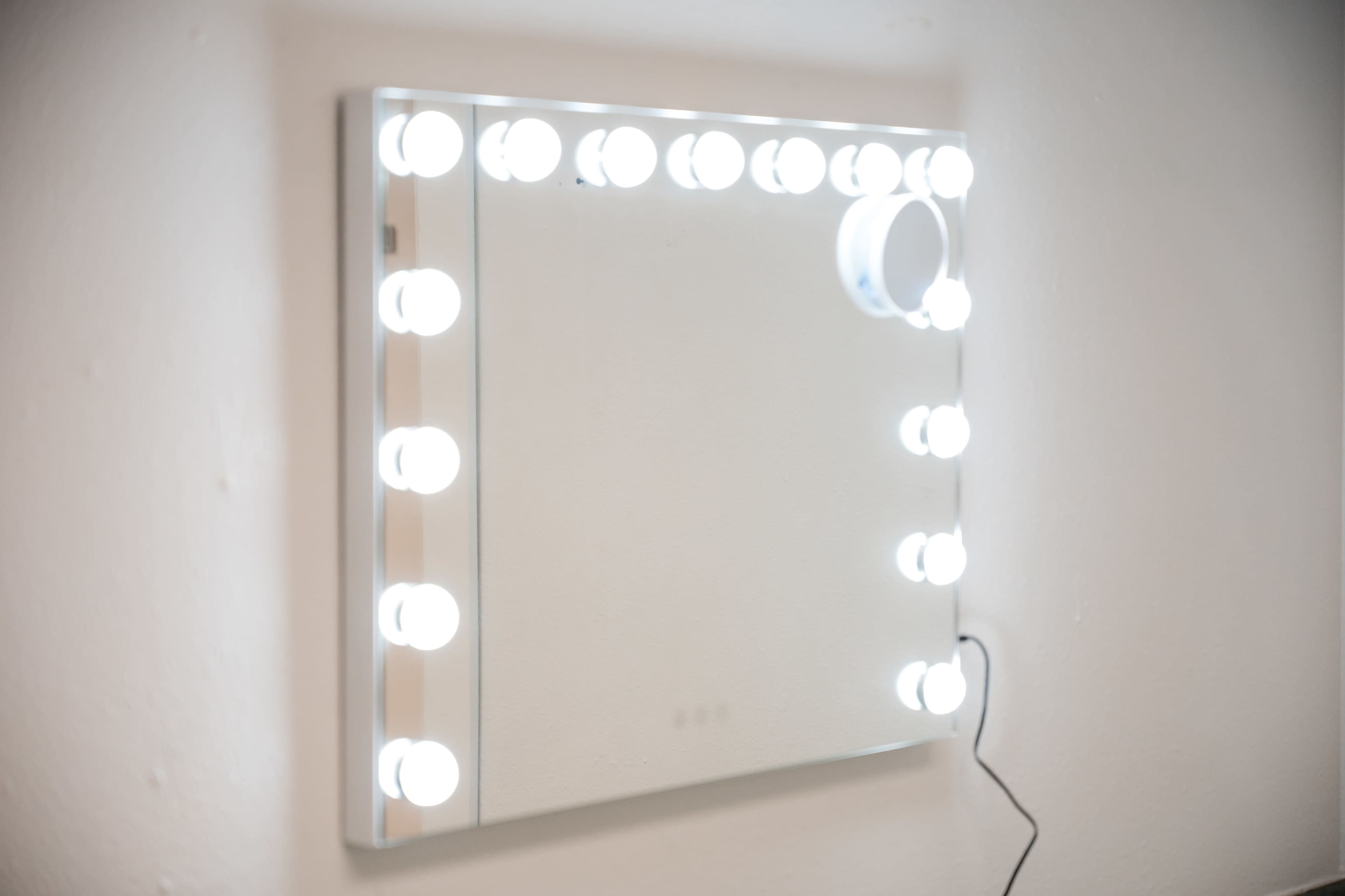 A square mirror with bright bulbs arranged around its edges is mounted on a wall.