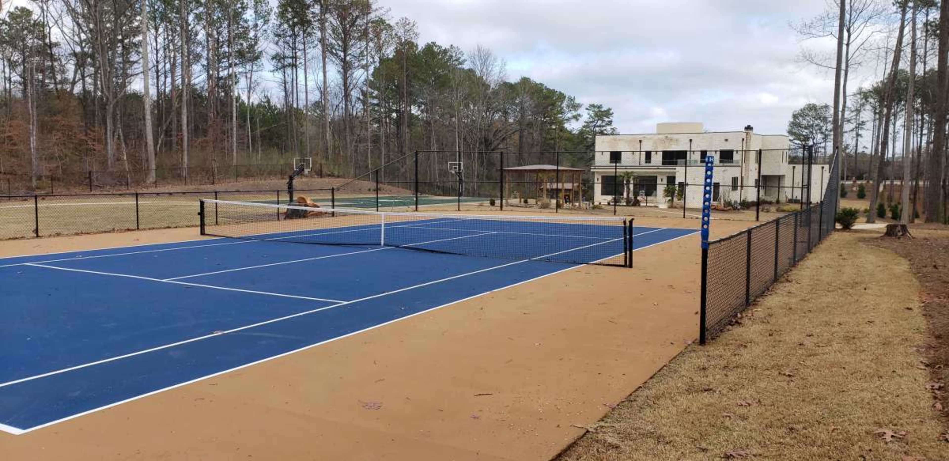 A well-maintained tennis court with a blue surface is positioned in front of a modern building surrounded by tall trees.
