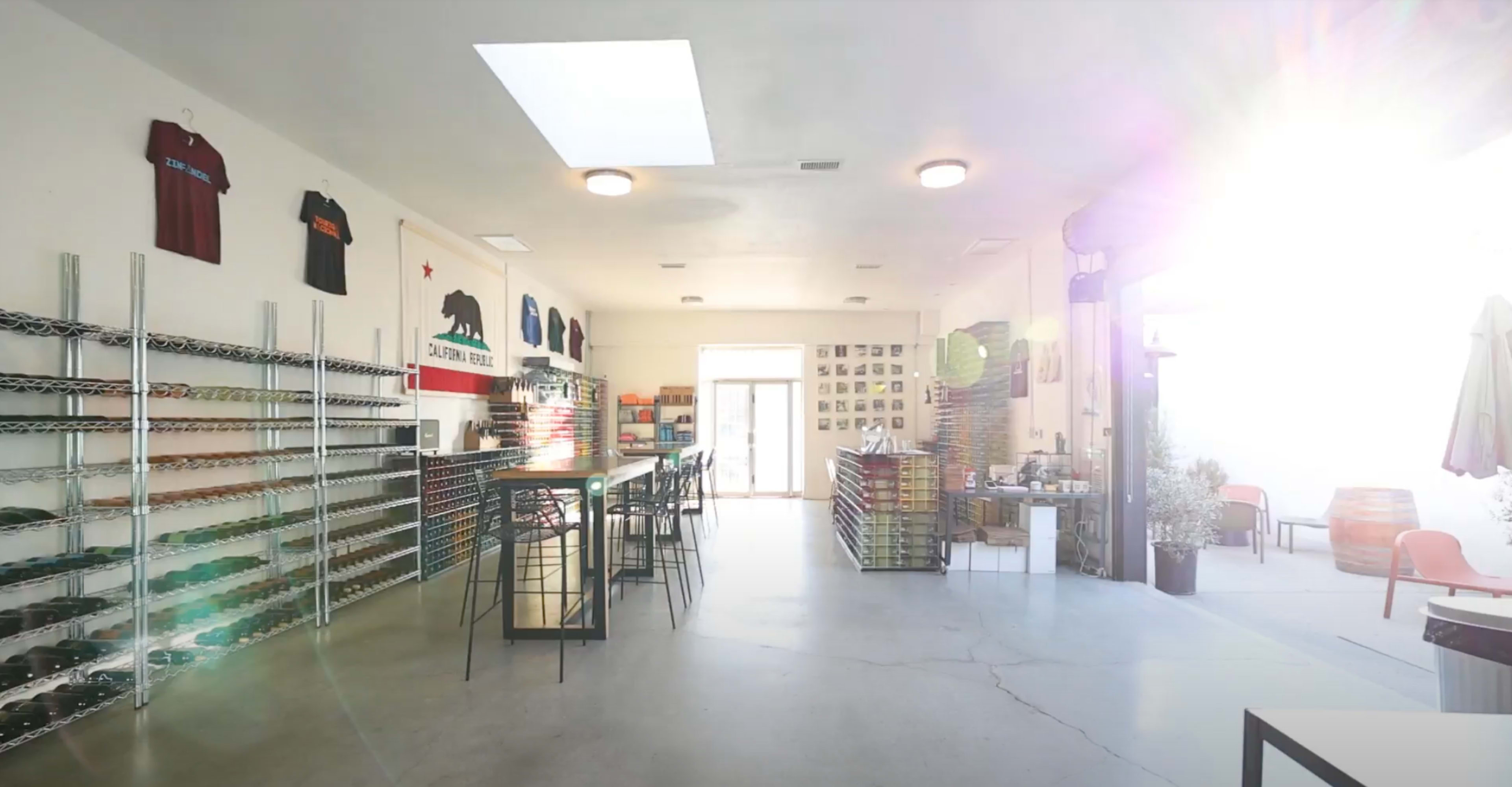 A brightly lit indoor space featuring metal shelving filled with various items, a long bar with stools, and a few tables and chairs.