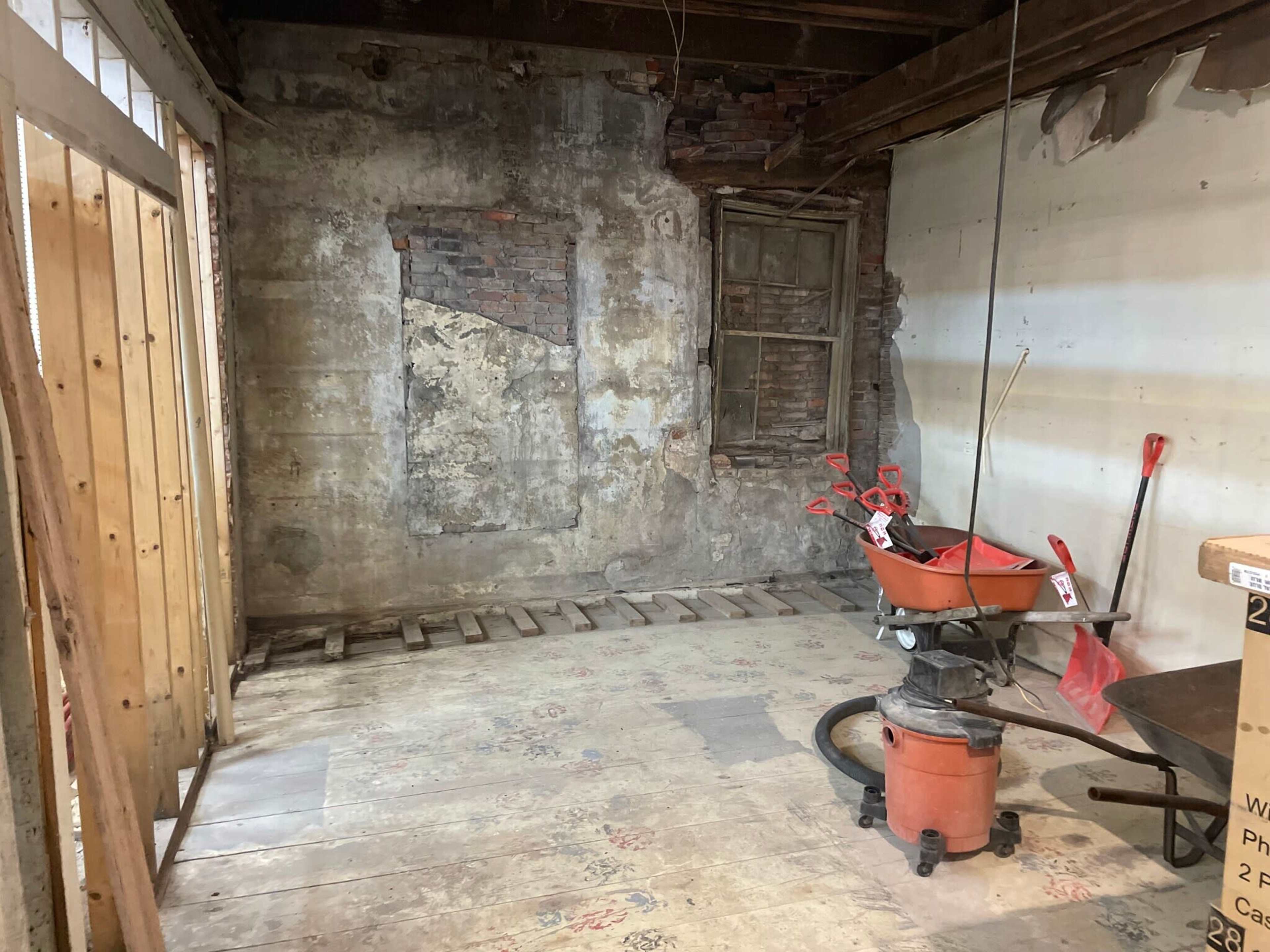 The image shows an unfinished room with exposed brick walls, a dusty floor, and construction materials, including a vacuum and shovels.