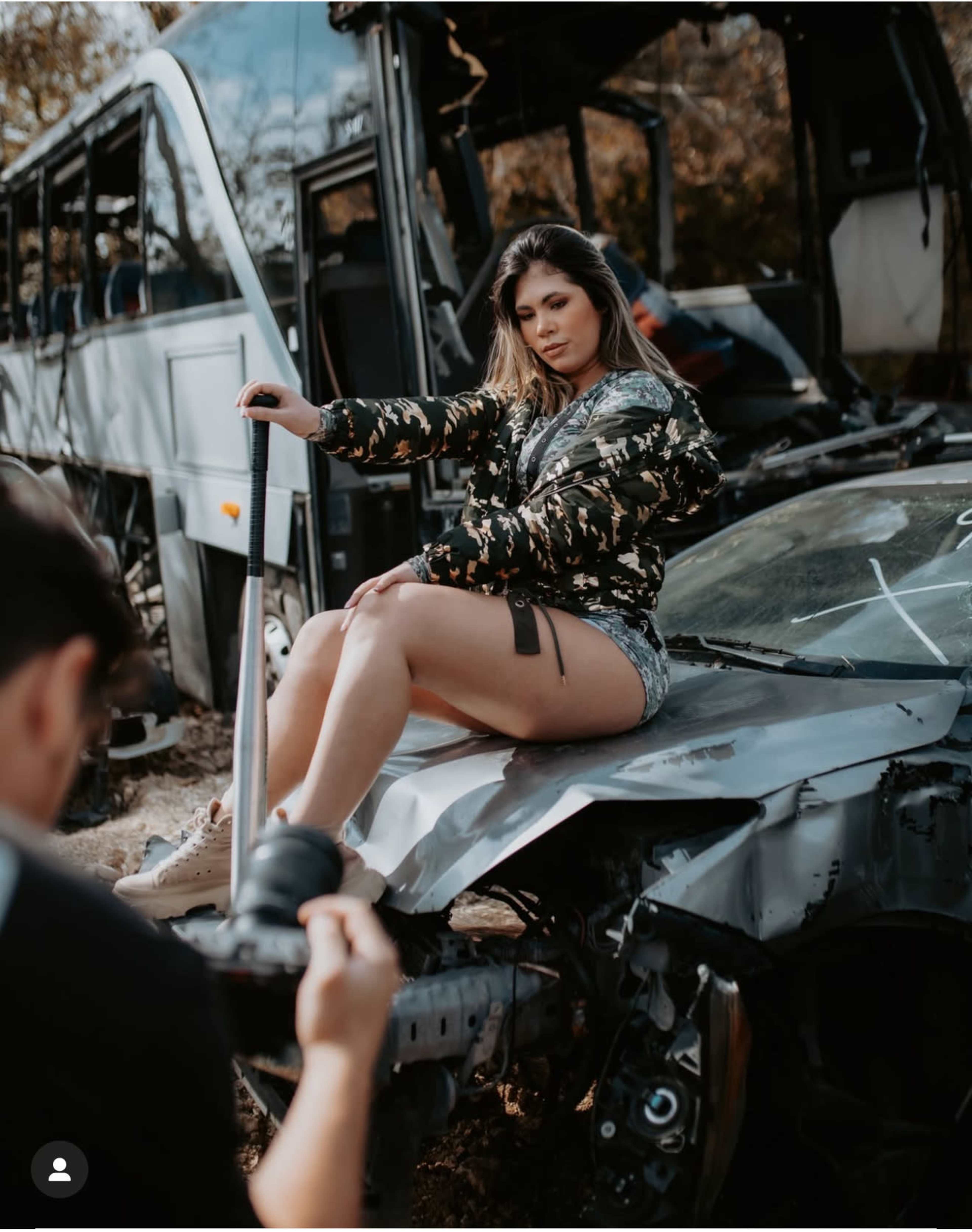A person poses on the hood of a damaged car in an area with abandoned vehicles.