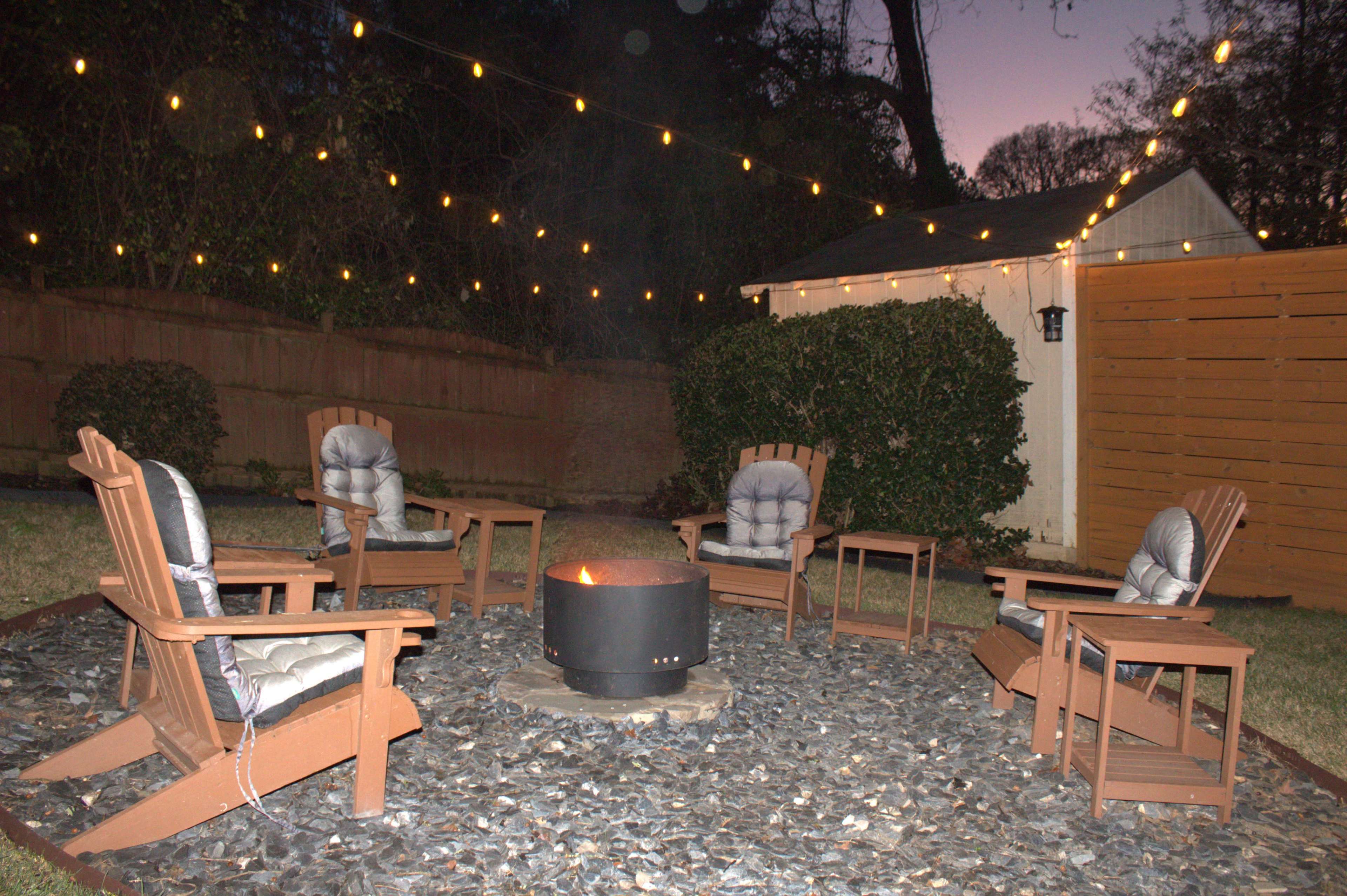 A cozy outdoor fire pit is surrounded by five wooden lounge chairs and adorned with string lights overhead, set against a backdrop of a shed and greenery.