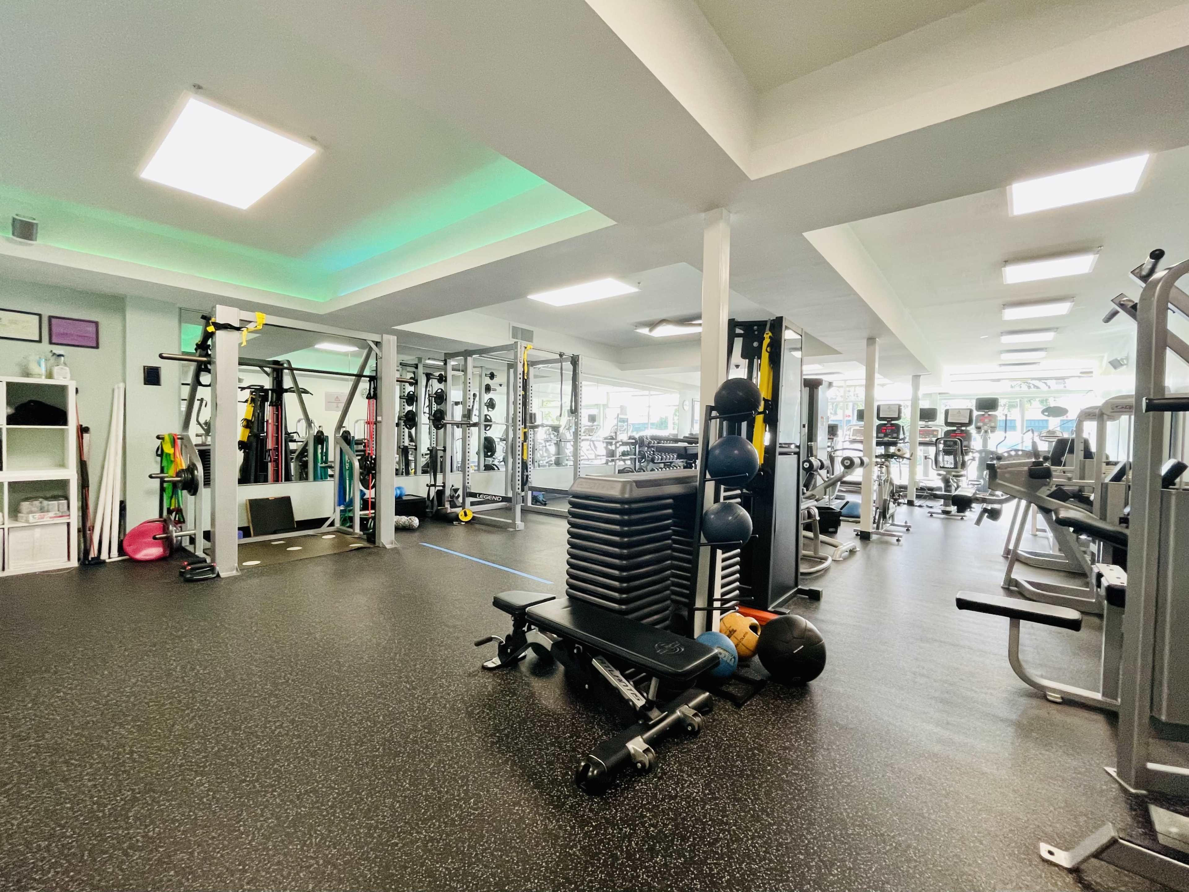 The image shows a well-equipped gym featuring various exercise machines, weights, and workout stations arranged across a spacious, brightly lit area.