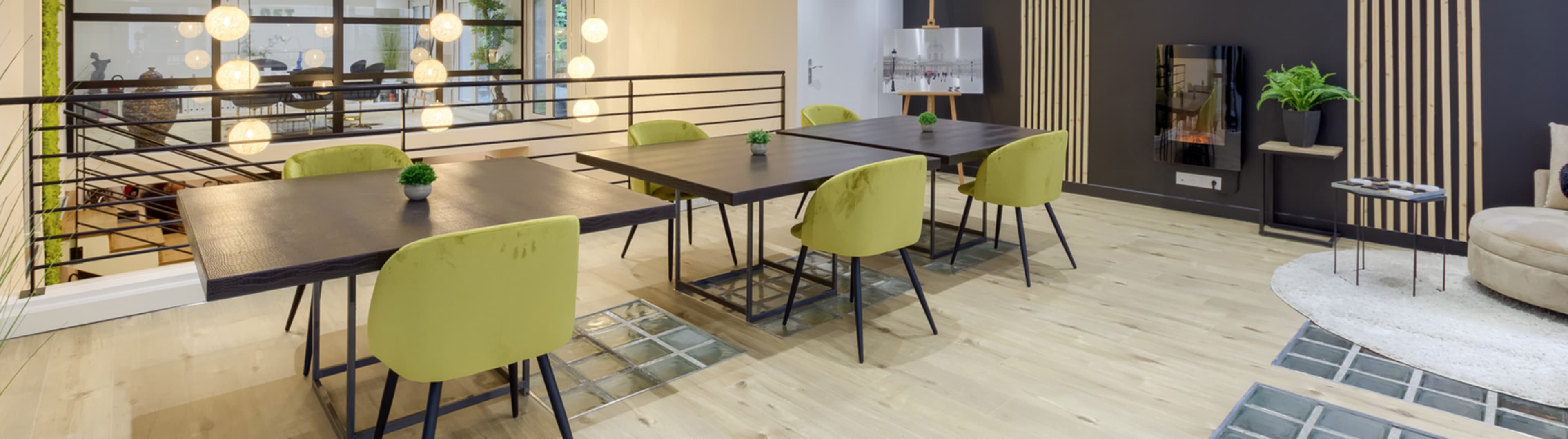 The image shows a modern interior with several tables and green chairs, illuminated by hanging light fixtures.