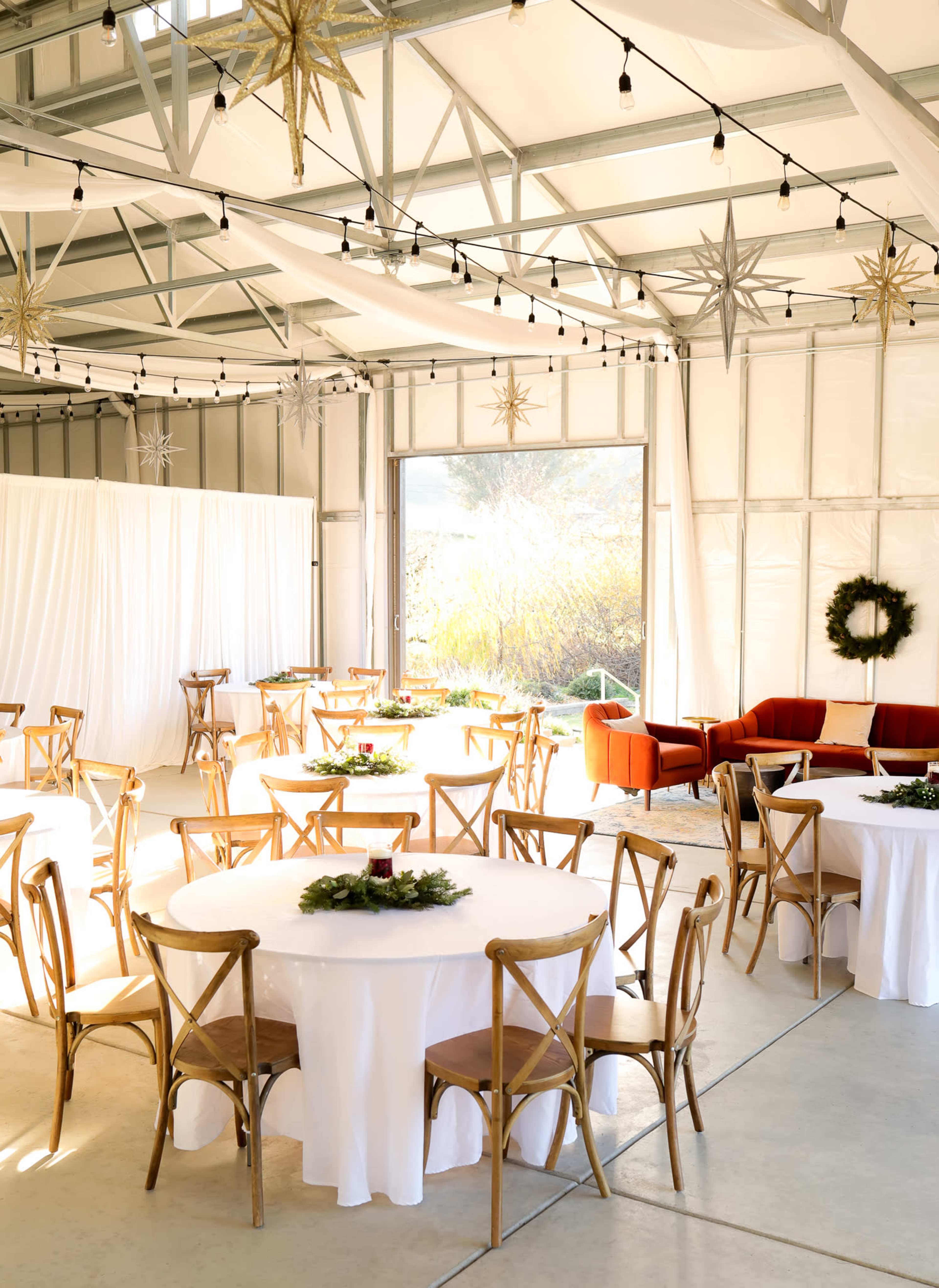 A spacious event space features round tables covered with white tablecloths, surrounded by wooden chairs, and a lounge area with a red sofa near a large open doorway.