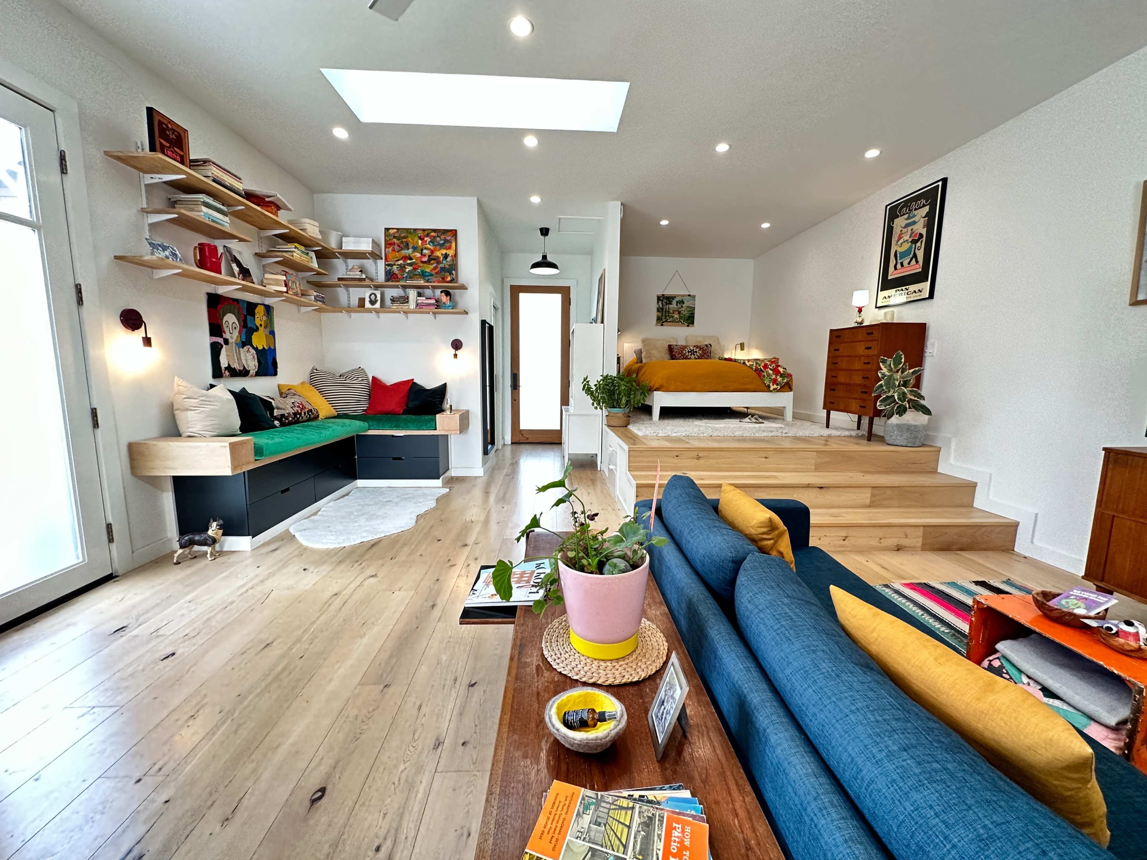 A modern interior space features a living area with a blue sofa, a wooden coffee table, and a bed on an elevated platform, surrounded by decorative shelves and art on the walls.