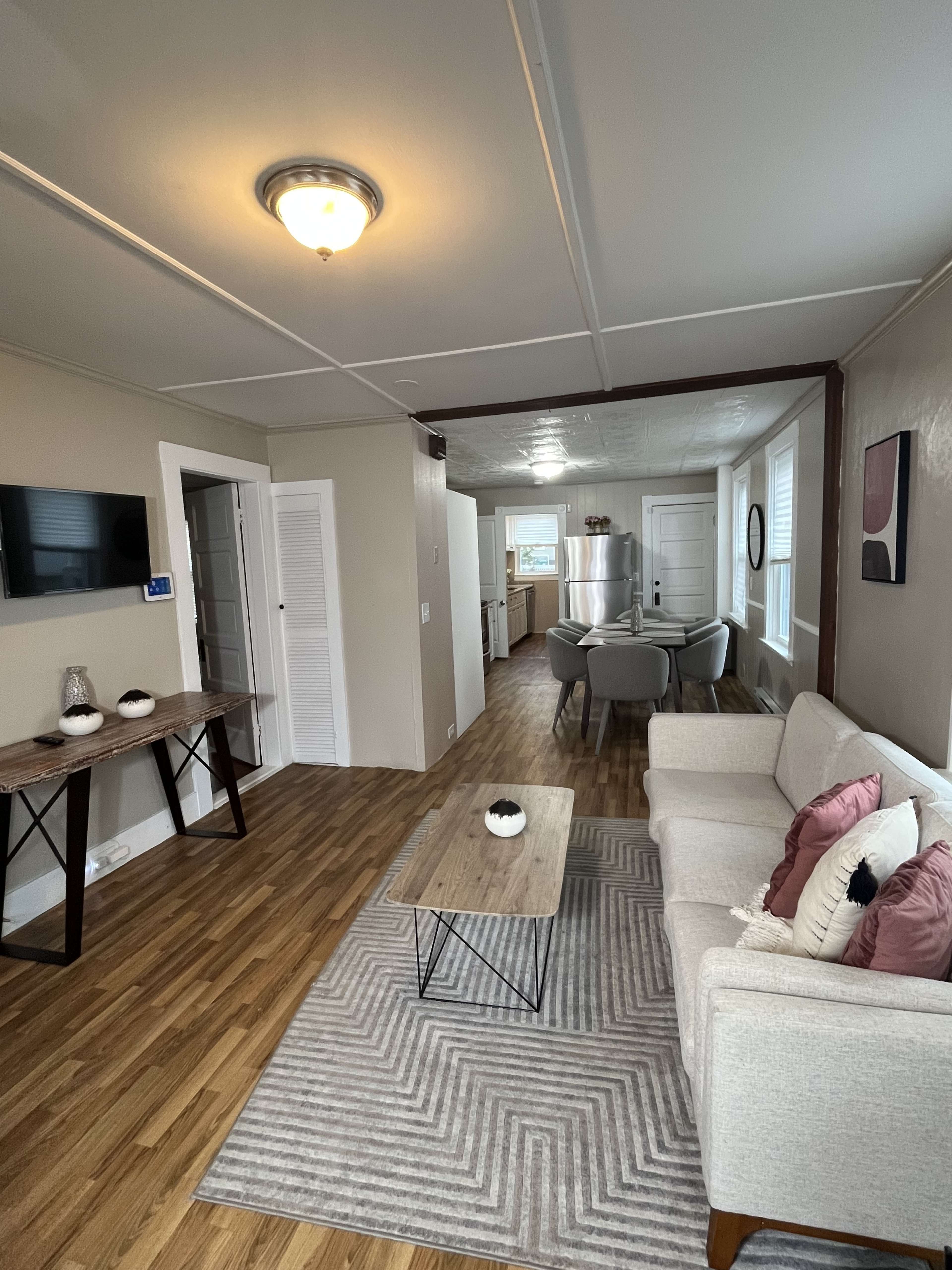 The image shows a cozy living area with a light-colored couch, a patterned rug, a coffee table, and a view into a kitchen space at the end of the room.